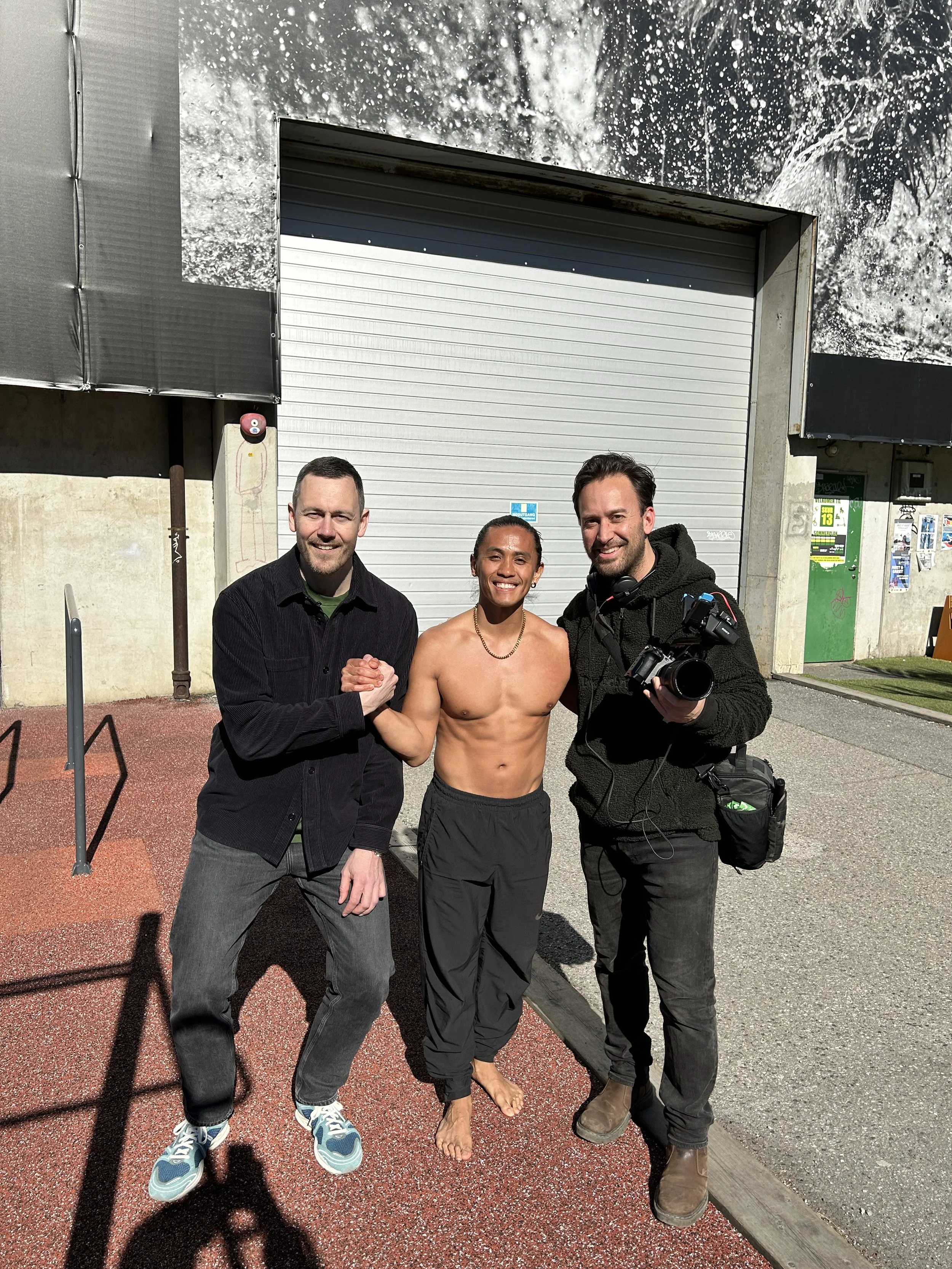 Three men and one woman standing together outdoors, posing for a photo in front of a closed gray garage door. The woman is barefoot, shirtless, smiling, and shaking hands with one of the men. The man on the right is holding a camera. The group is standing on a red textured surface, and there is a building with posters and signs on the wall behind them.
