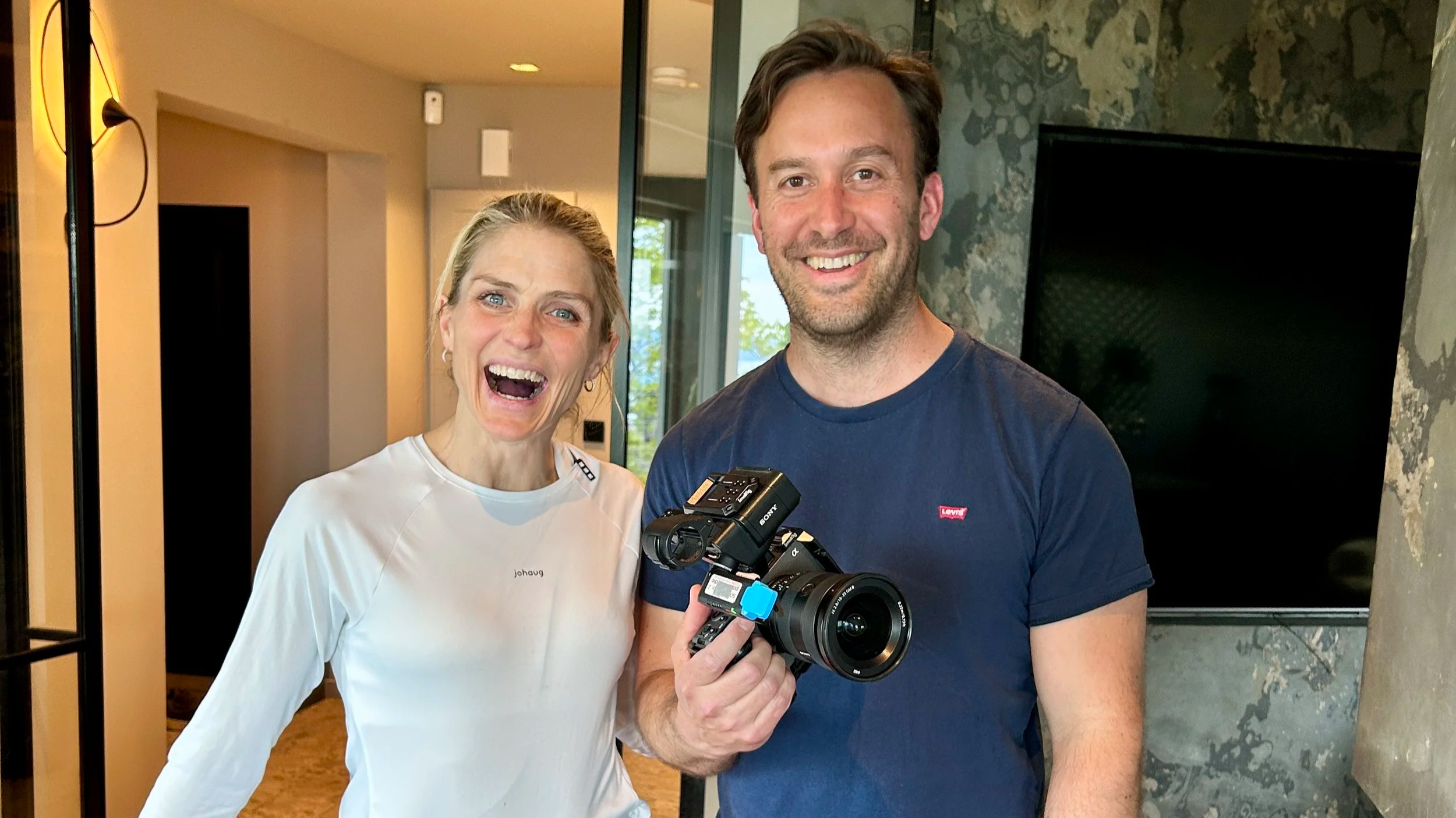 A woman and man smiling, with the man holding a professional video camera. They are indoors near a television and a window.