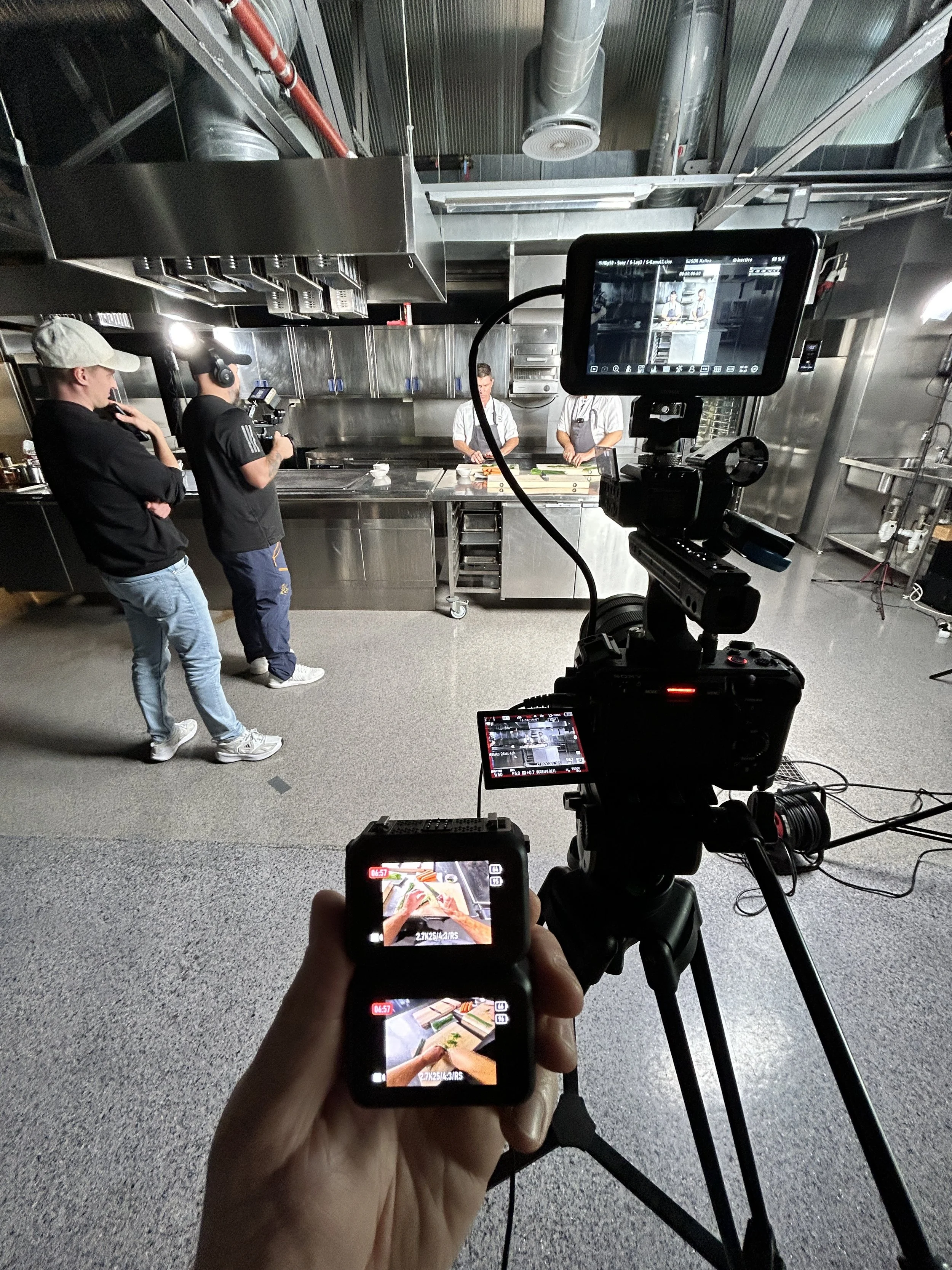 A behind-the-scenes view of a video shoot in a kitchen studio. Two men wearing black shirts and hats record two chefs preparing food at a stainless steel counter. A professional camera on a tripod captures the scene, and the footage is monitored on a large display.