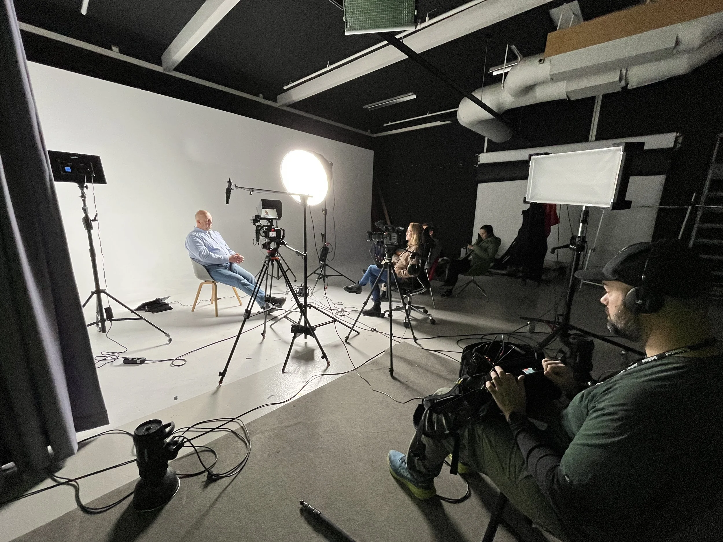 People sitting in chairs during a video recording or interview in a studio with lighting and camera equipment.