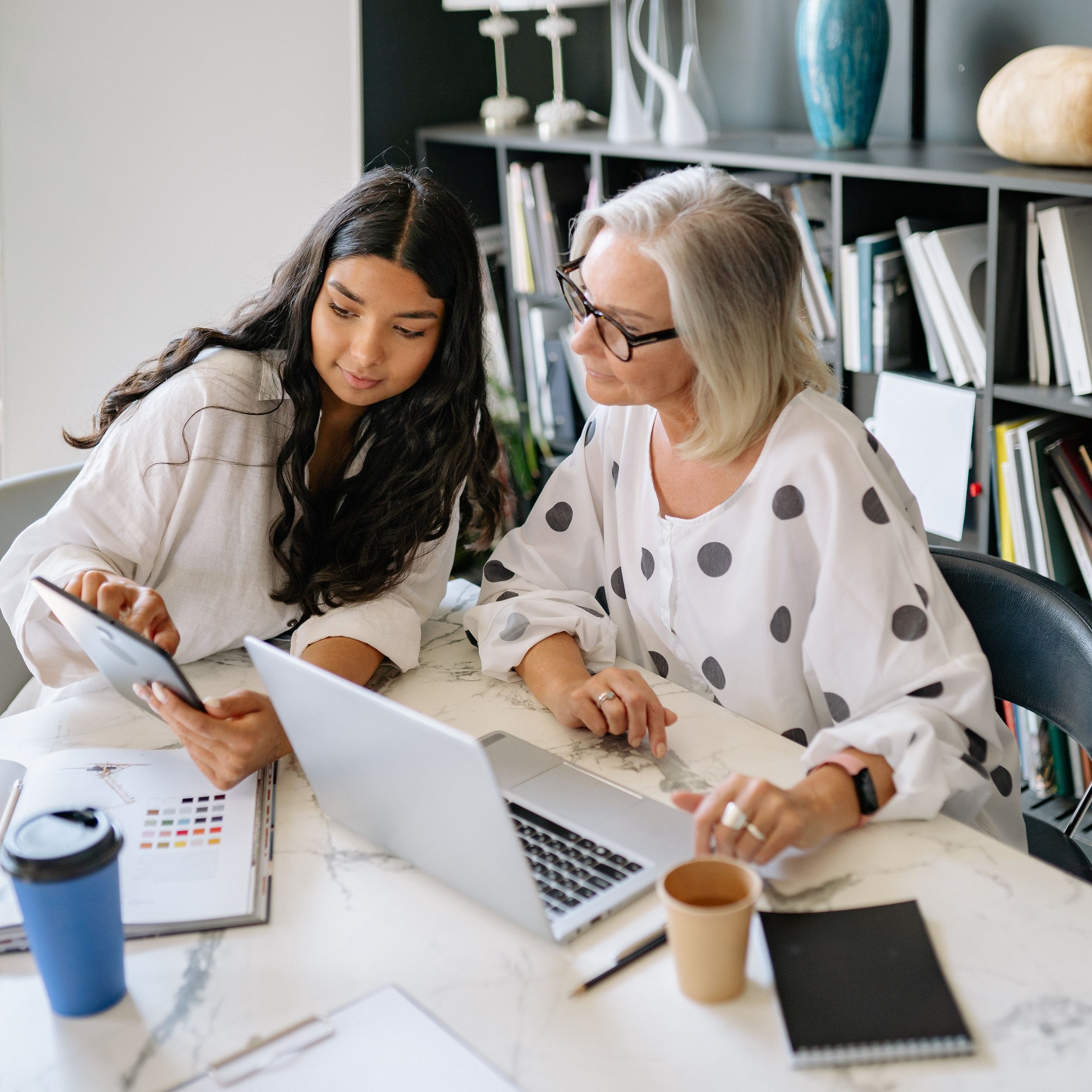 Two women sitting at a table working together with laptops, papers, and coffee cups, in a modern office with a bookshelf and decorative items in the background.