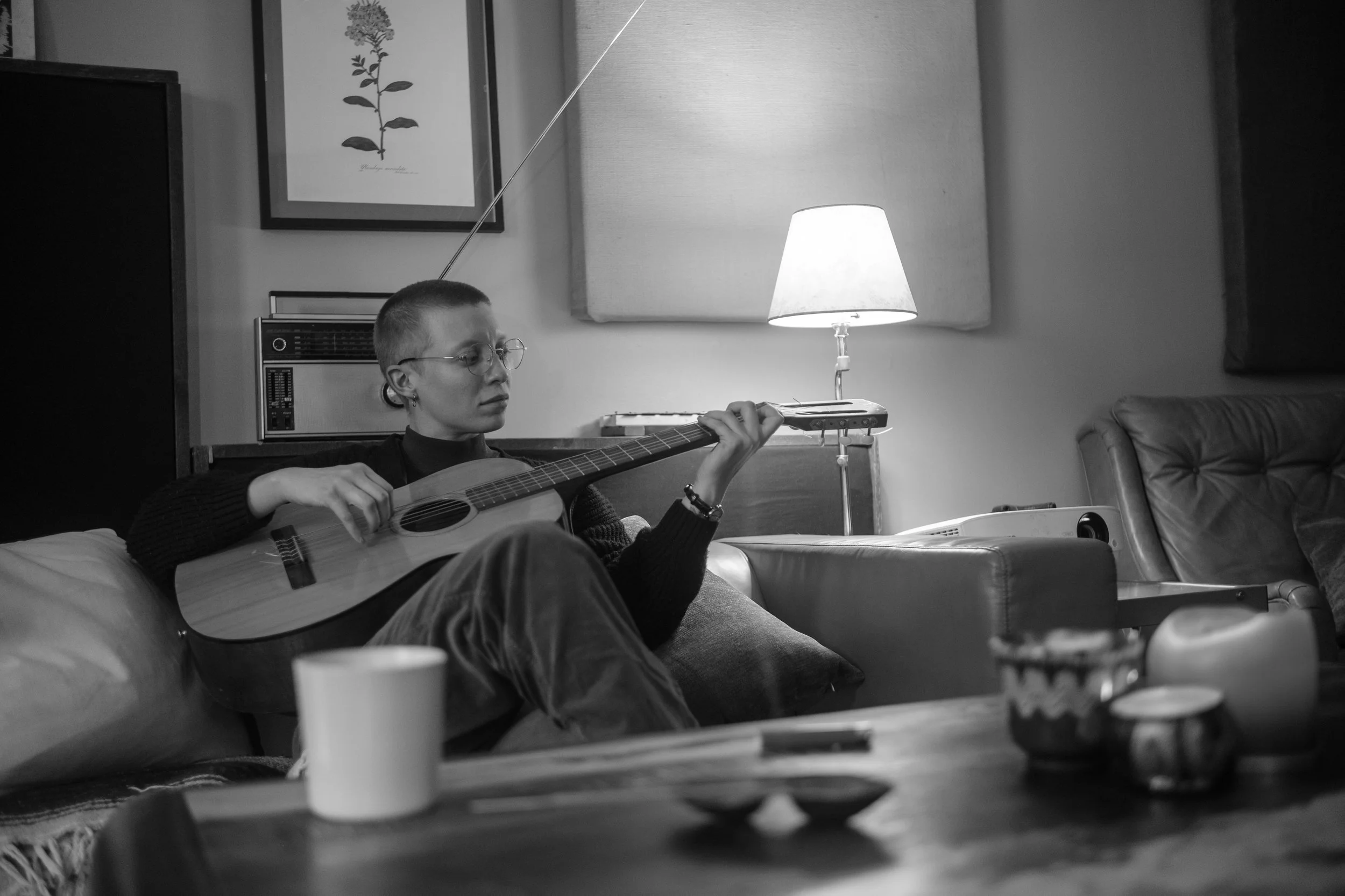 A person playing an acoustic guitar while sitting on a sofa in a living room, with a lamp on a side table, a framed picture on the wall, and a heater behind them.