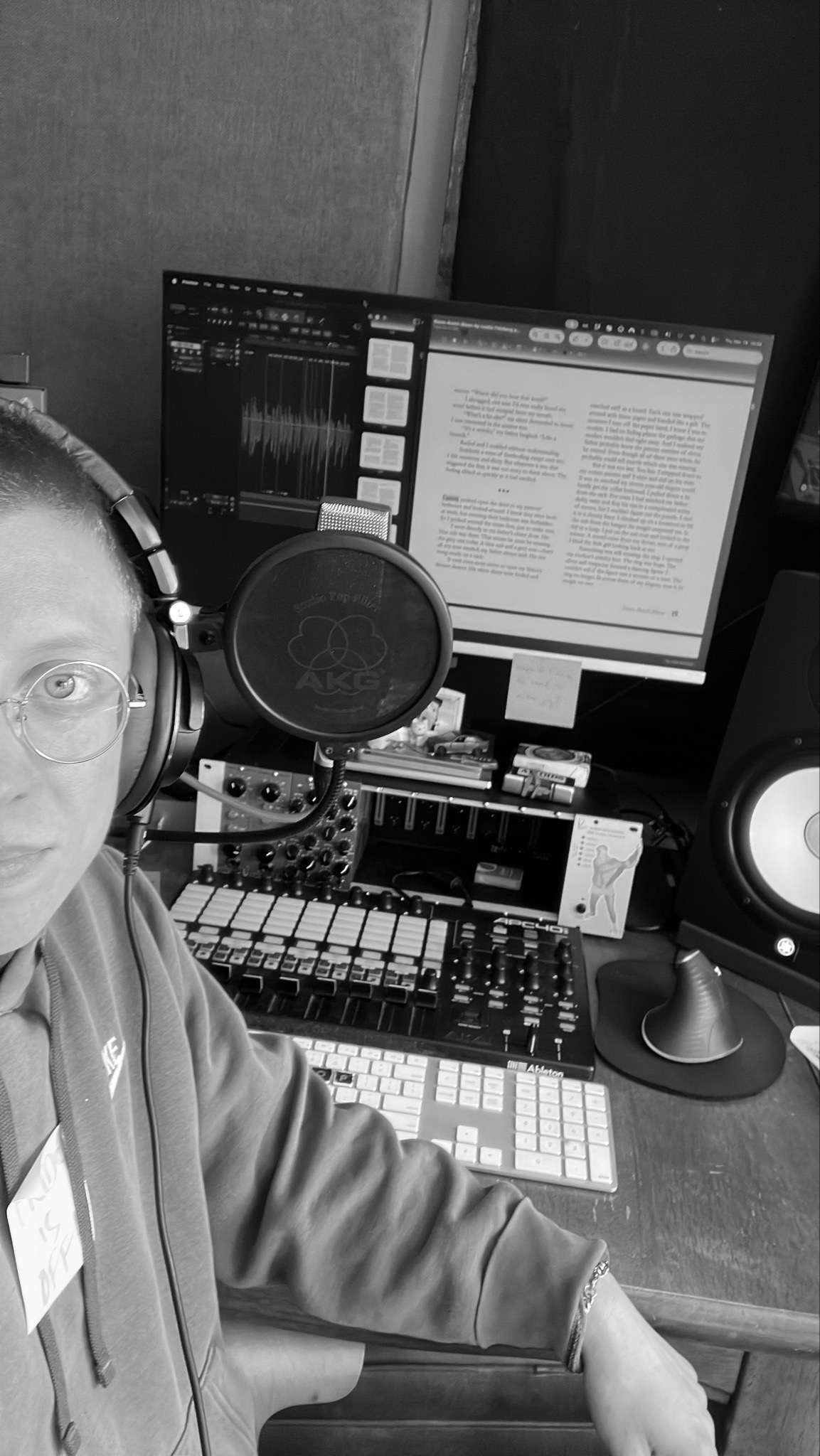 A person wearing glasses and headphones in a music studio surrounded by music production equipment, including monitors, a keyboard, a microphone, and a computer screen with audio editing and text documents.