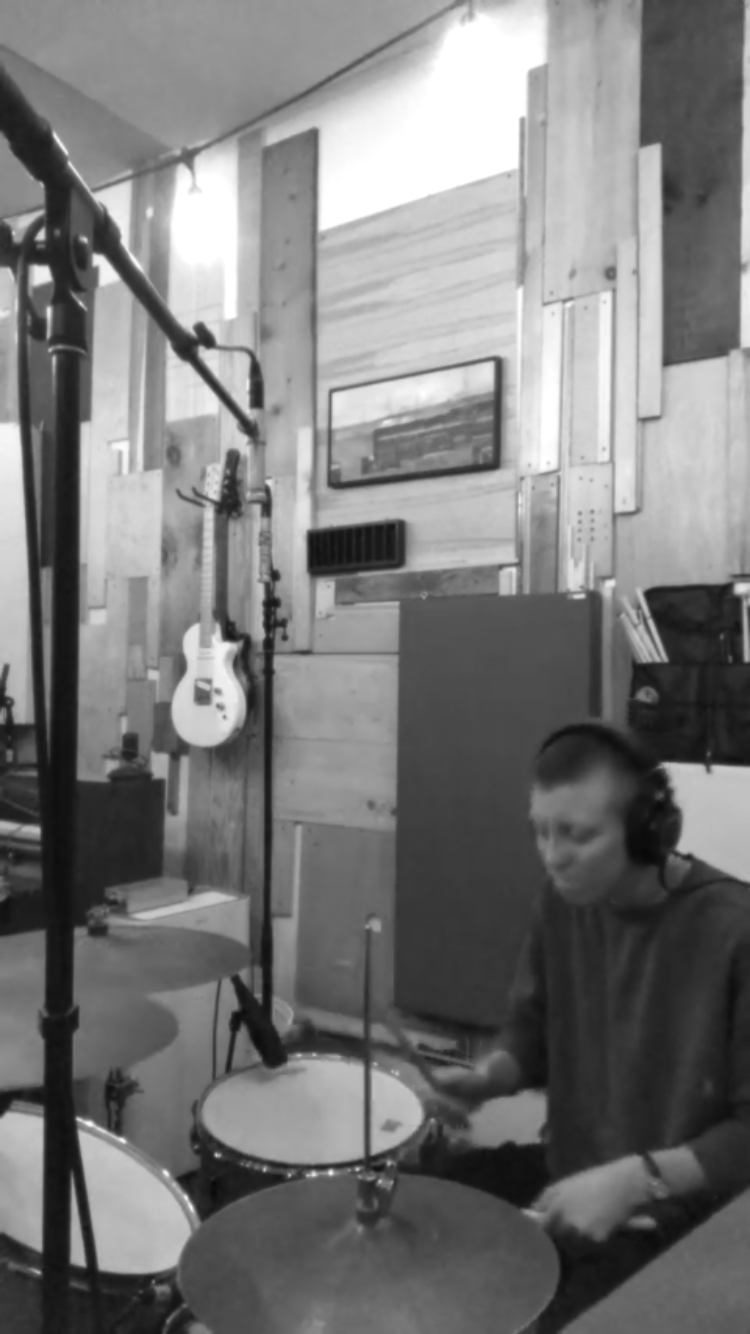 A person wearing headphones playing drums in a recording studio with wooden walls, a framed picture, and a guitar hanging on the wall.