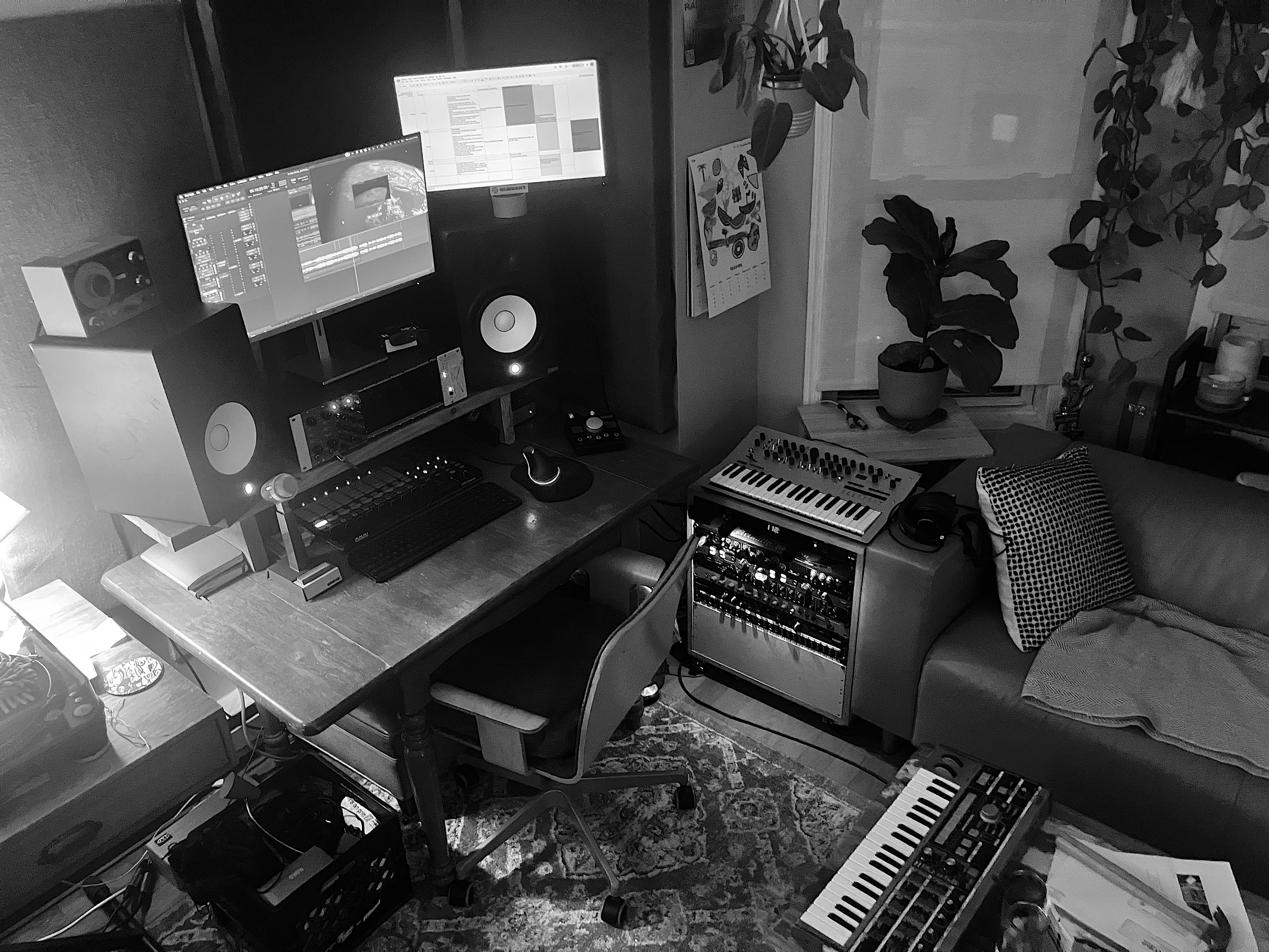 A music studio setup with two computer monitors displaying editing software, speakers, a computer keyboard, a small MIDI keyboard, a mixer, and various other electronic music equipment arranged on a wooden desk. There are also potted plants, a calendar, and a patterned cushion in the room.