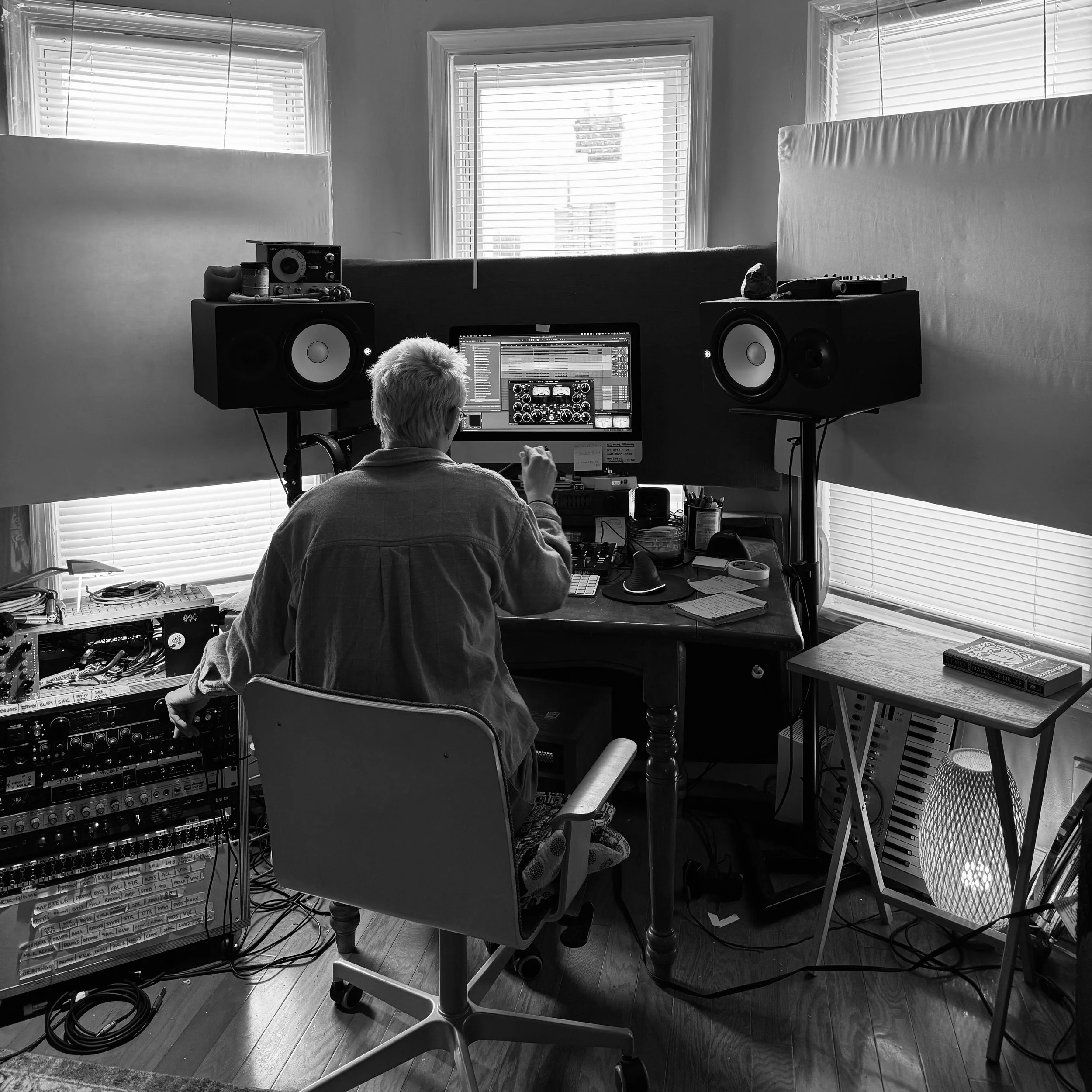 A person is working on music production at a desk with a computer, studio speakers, and audio equipment. The room has large windows with blinds, and there are various cables, devices, and books scattered around.