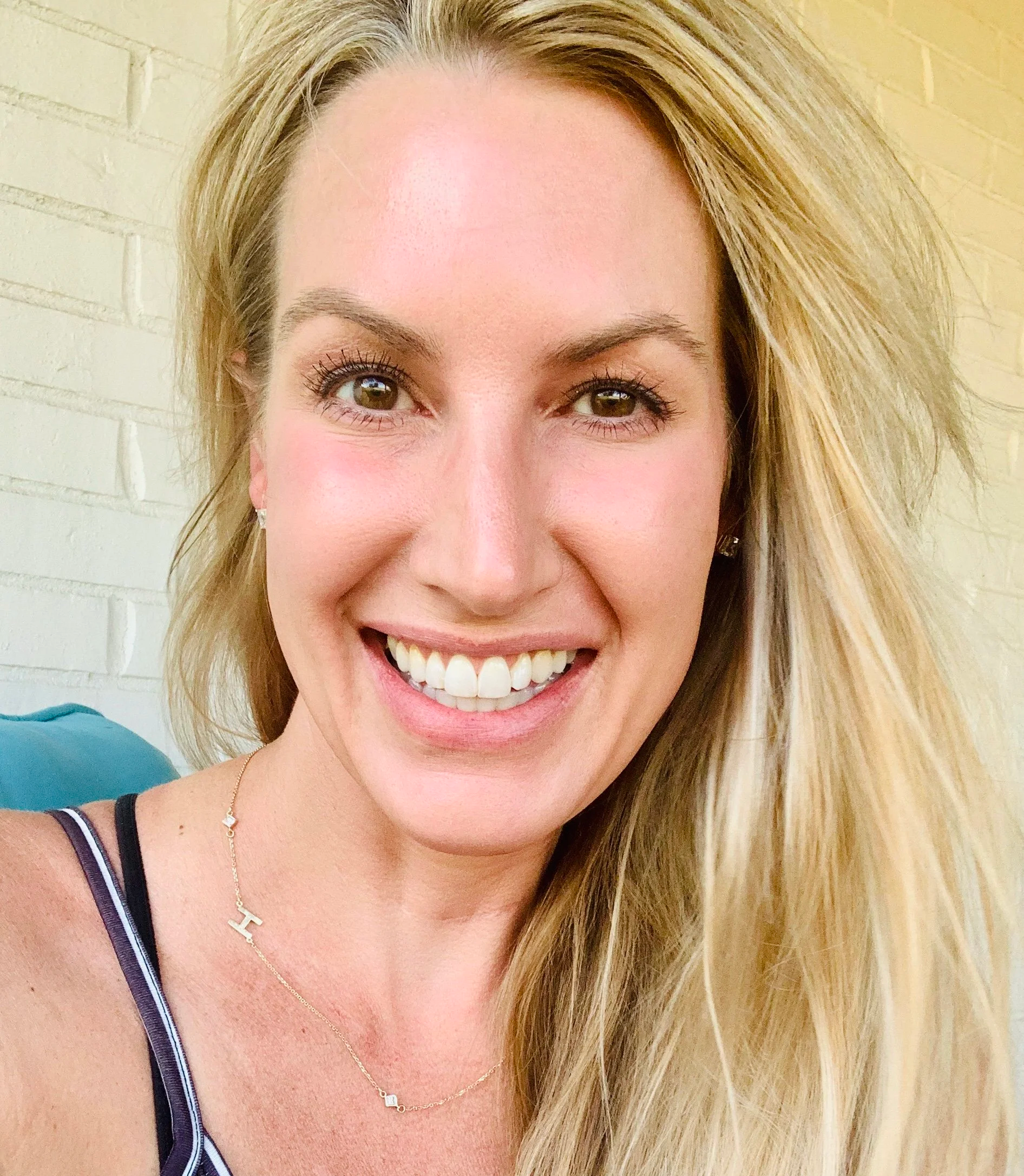 Close-up of a woman smiling, with long blonde hair, green eyes, wearing a necklace and a black and white striped top, standing against a light-colored brick wall.