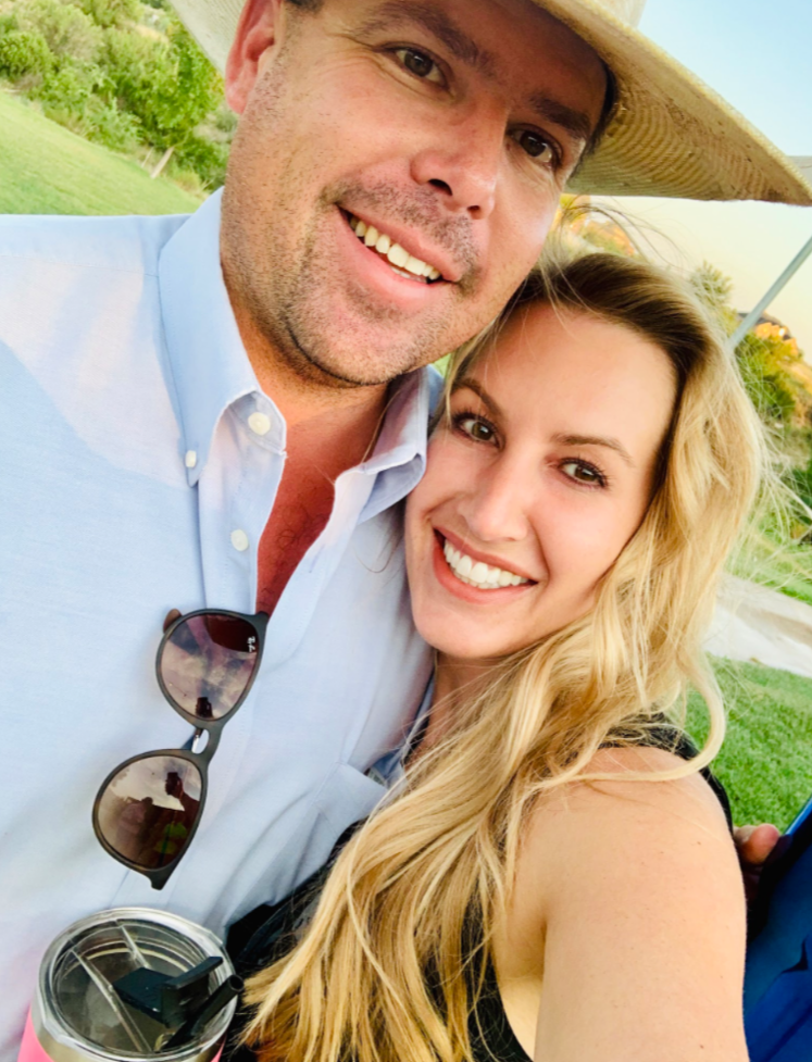 A smiling man and woman taking a selfie outdoors on a sunny day, with lush green trees and grass in the background. The man is wearing a light blue button-up shirt and a wide-brimmed hat, with sunglasses hanging from his shirt. The woman has long blonde hair and is wearing a black top, holding a drink with a straw.