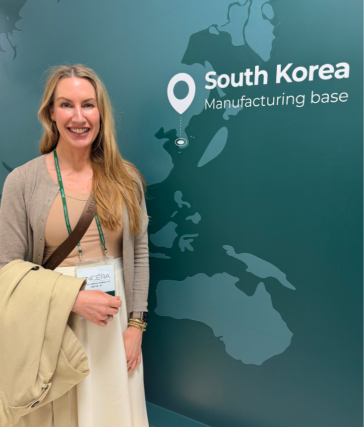 A smiling woman with long blonde hair standing next to a map of the world showing South Korea as a manufacturing base, at the AceCosm headquarters.