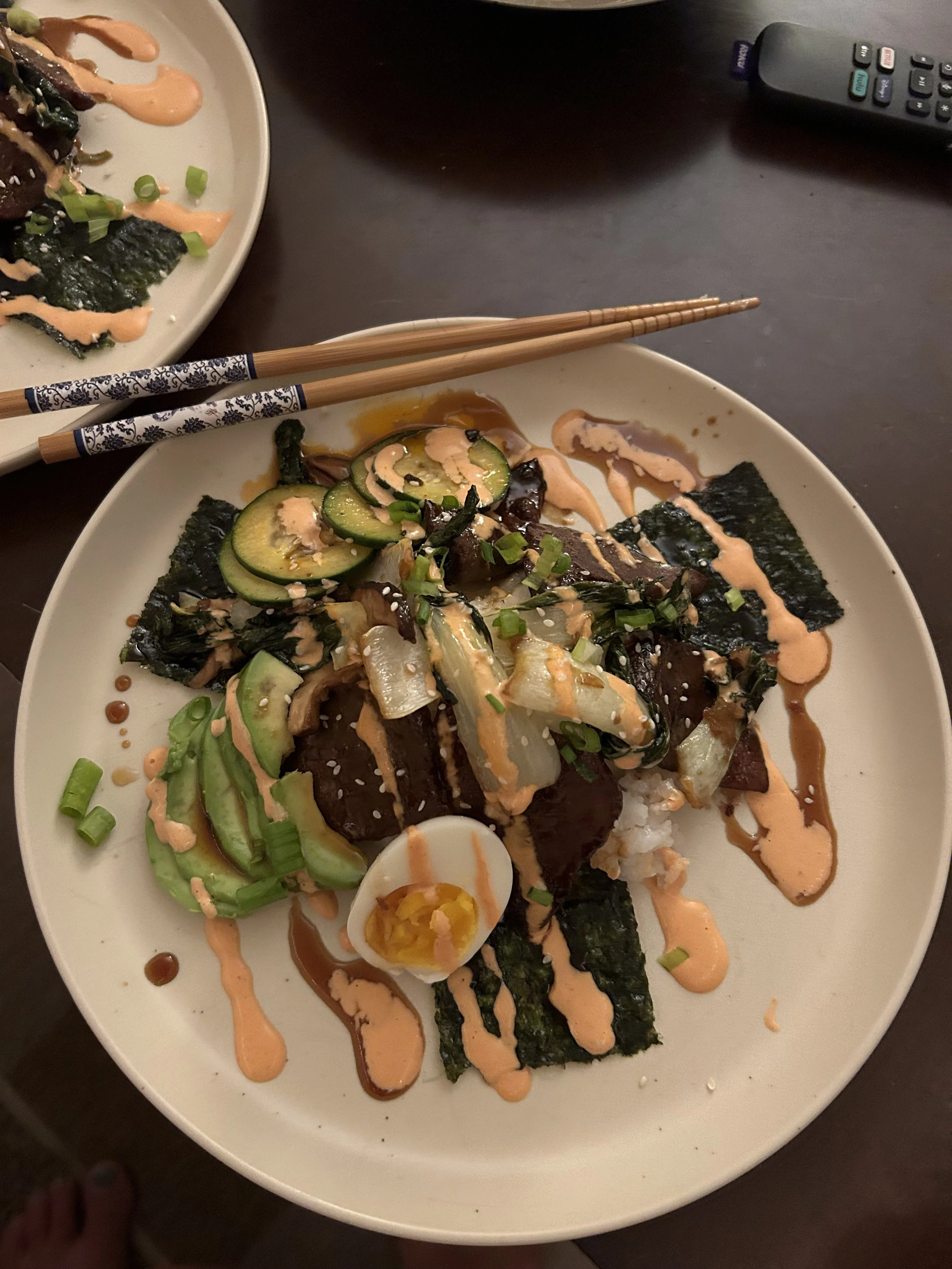 A white plate with sushi rolls topped with sliced cucumber, green onions, seaweed, and drizzled with orange sauce. There are also pieces of seaweed and a boiled egg cut in half on the plate. In the background, there is another plate with similar sush