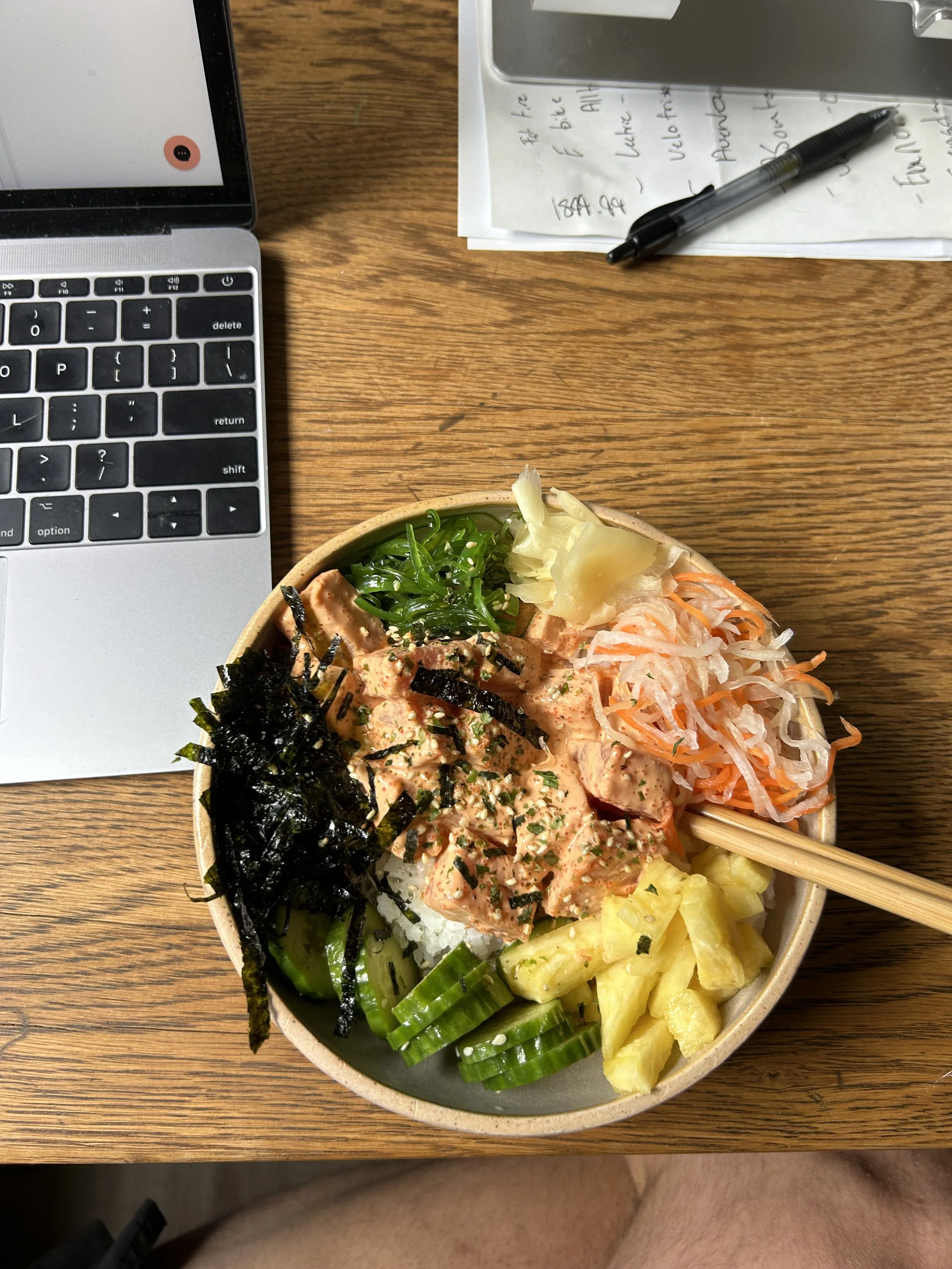 A bowl of poke with rice, chopped pineapple, shredded carrots, green onions, seaweed, and seasoned raw fish, placed on a wooden table.