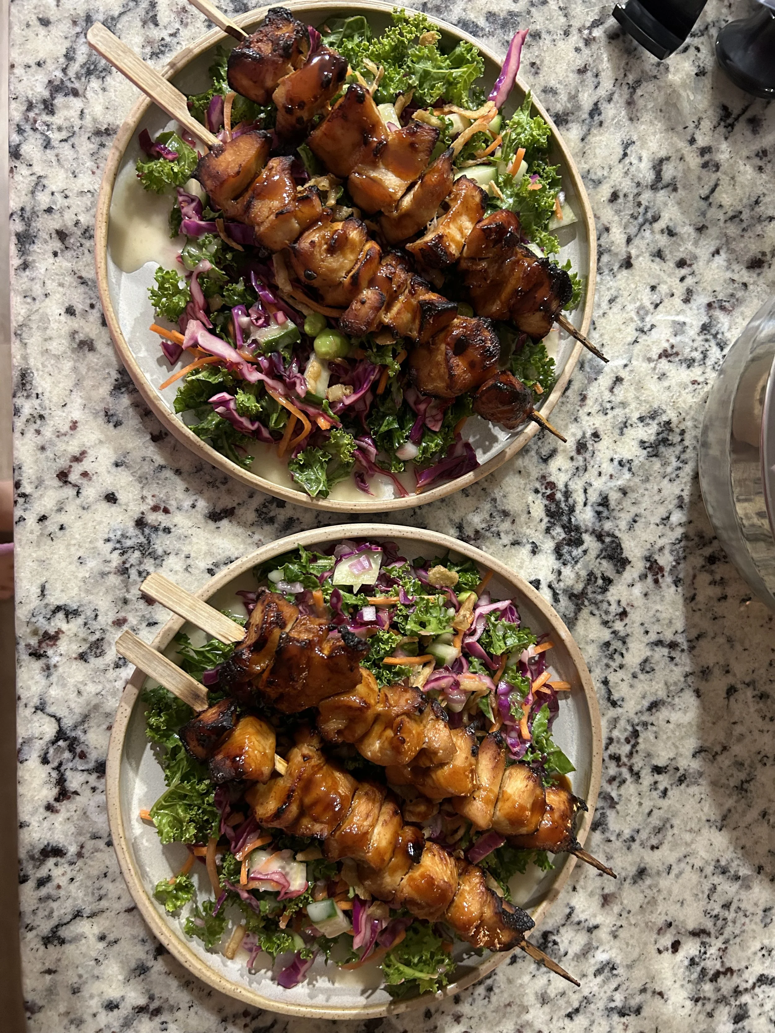 Two oval plates of grilled chicken skewers on a bed of colorful salad greens and vegetables, on a speckled granite countertop.