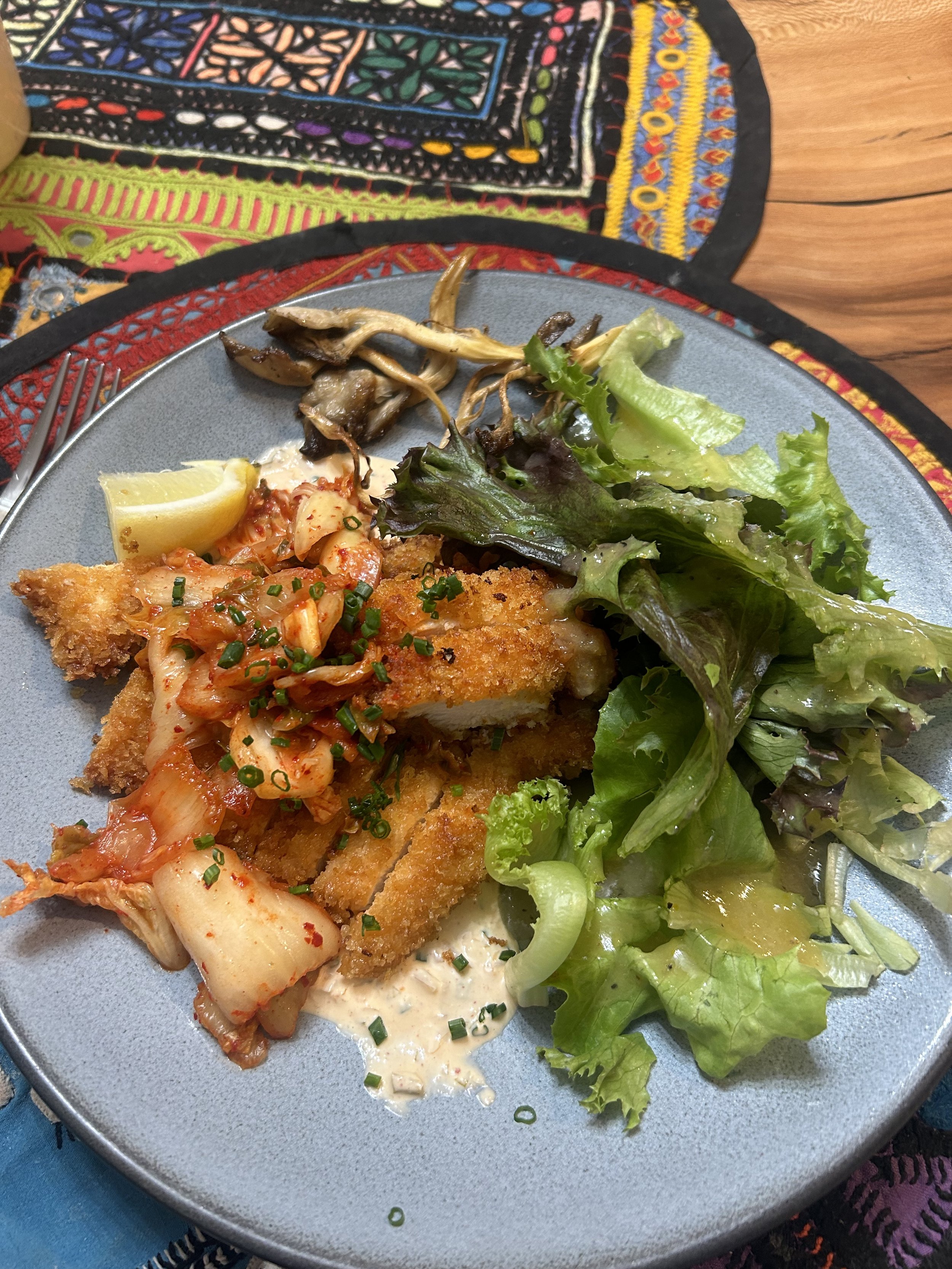 A plate of food with fried fish topped with kimchi, a lemon wedge, and green onion, next to a side salad with mixed greens, and cooked mushrooms, on a table with colorful embroidered fabric.