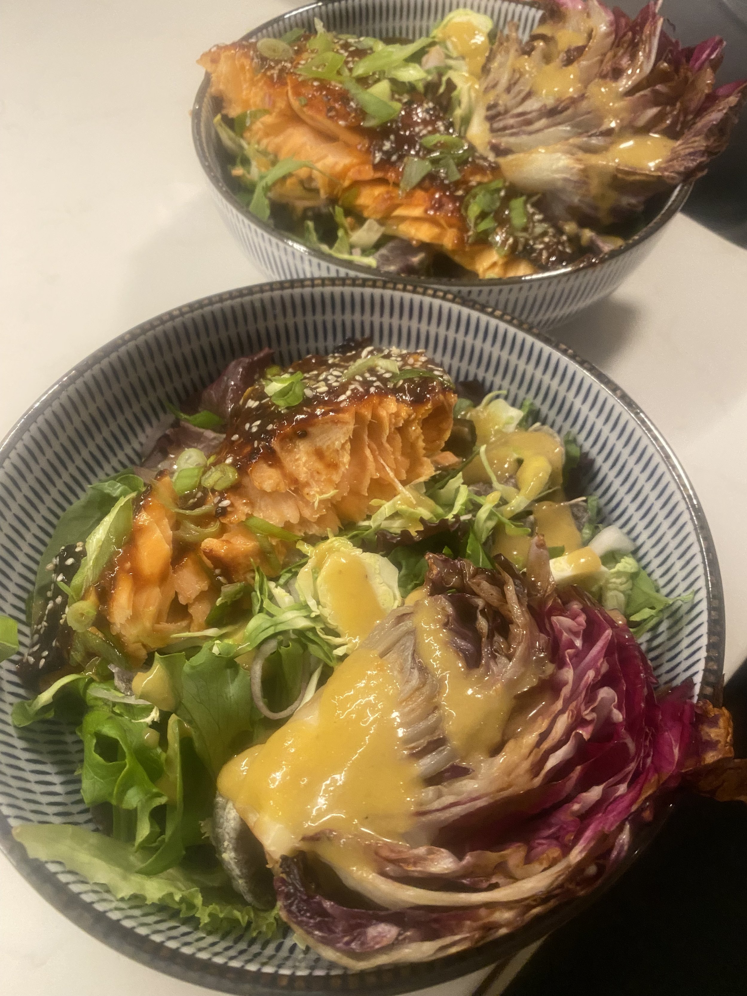 A bowl with a leafy green salad topped with grilled salmon, chopped green onions, sesame seeds, and a dark sauce. Another bowl contains a green salad with a grilled salmon fillet, chopped green onions, sesame seeds, and yellow dressing. Both bowls ha
