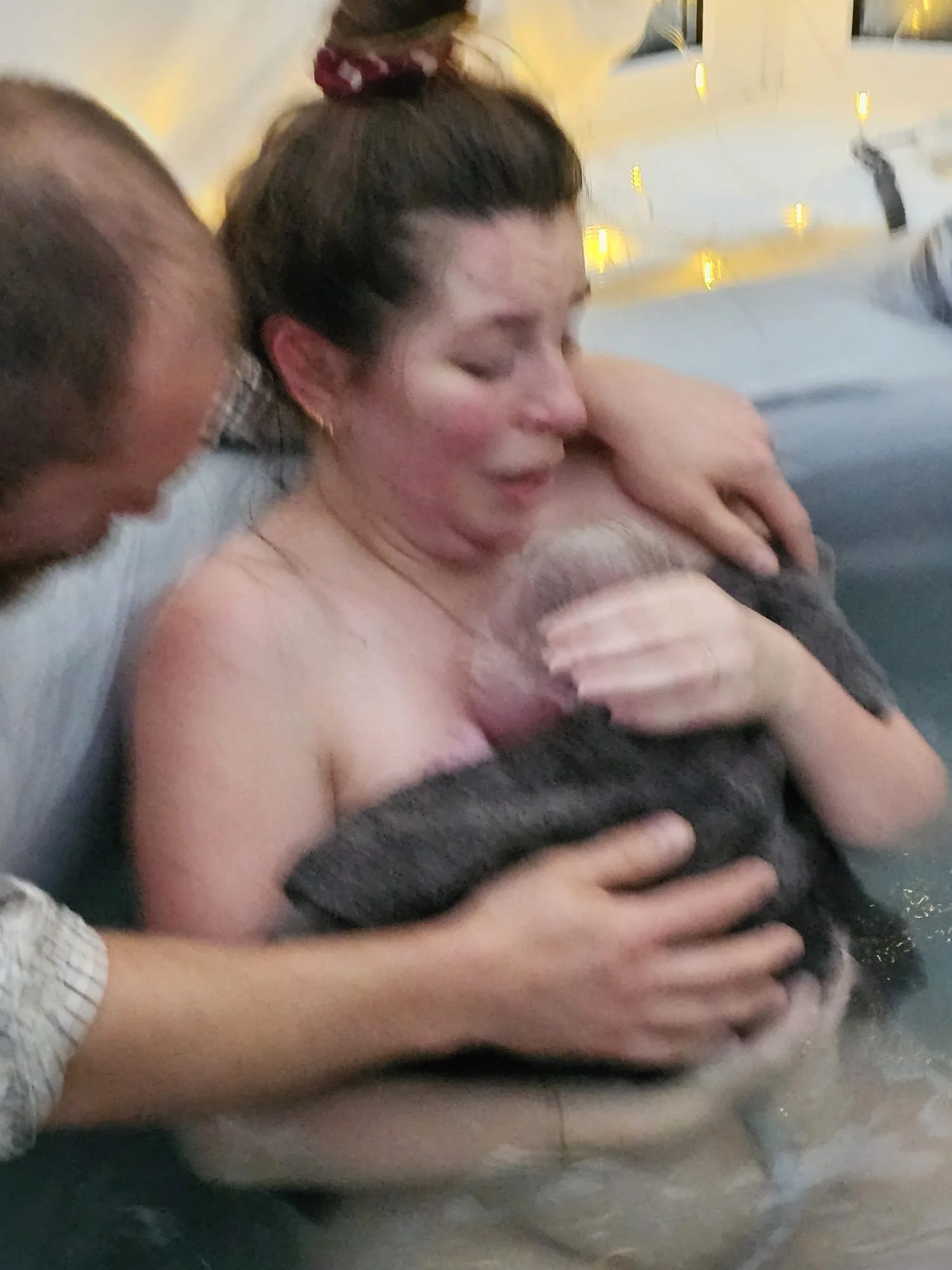 A woman with dark hair tied in a bun with a red hair tie, holding and cuddling a freshly born baby close to her chest, with her eyes closed in a tender moment.