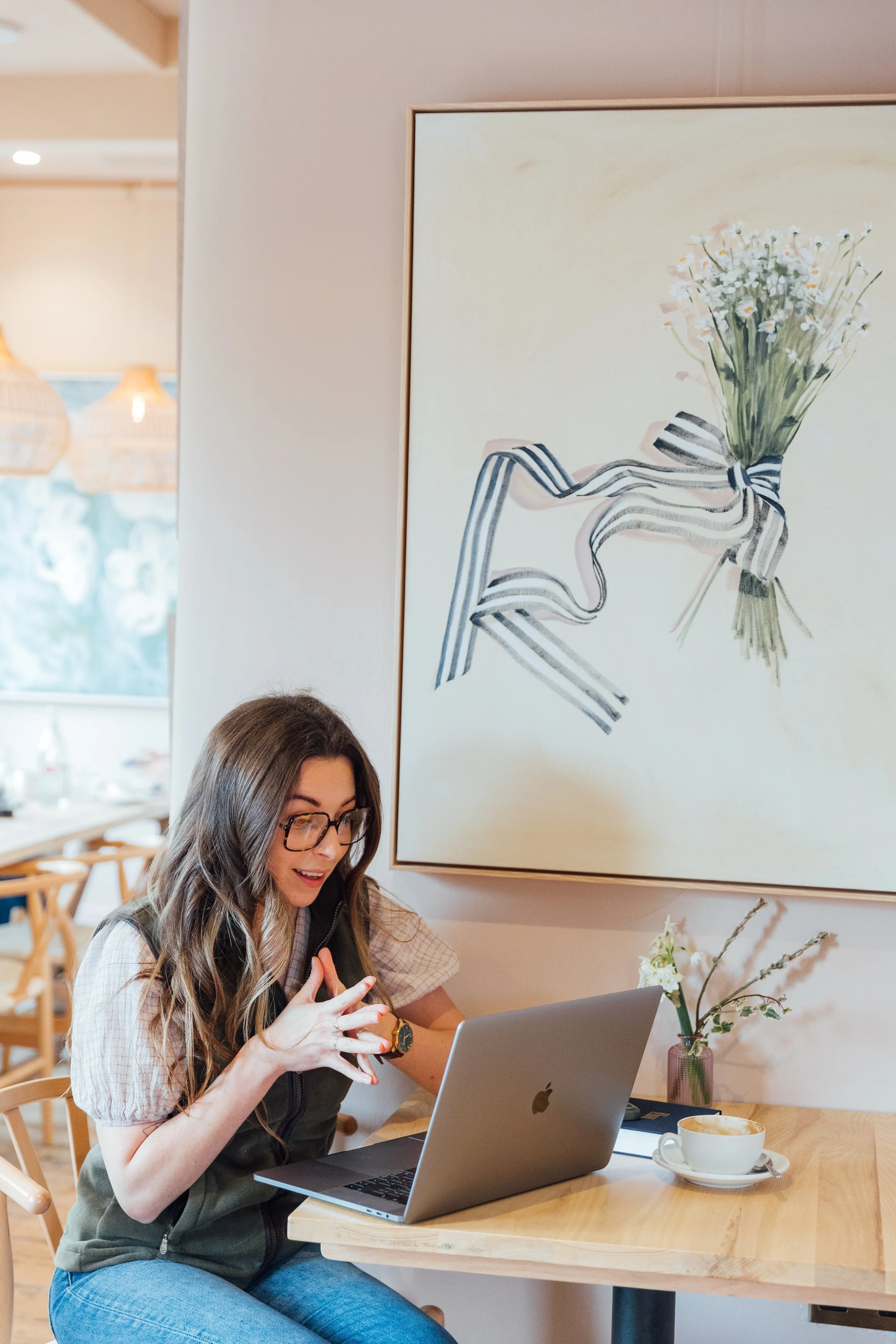 A woman with long brown hair and glasses sitting at a table in a cafe, looking at her phone, with a laptop in front of her and a white coffee cup on the table. There is a flower vase with white flowers on her right, and a large wall art of a bouquet of flowers tied with a striped ribbon hanging on the wall behind her.