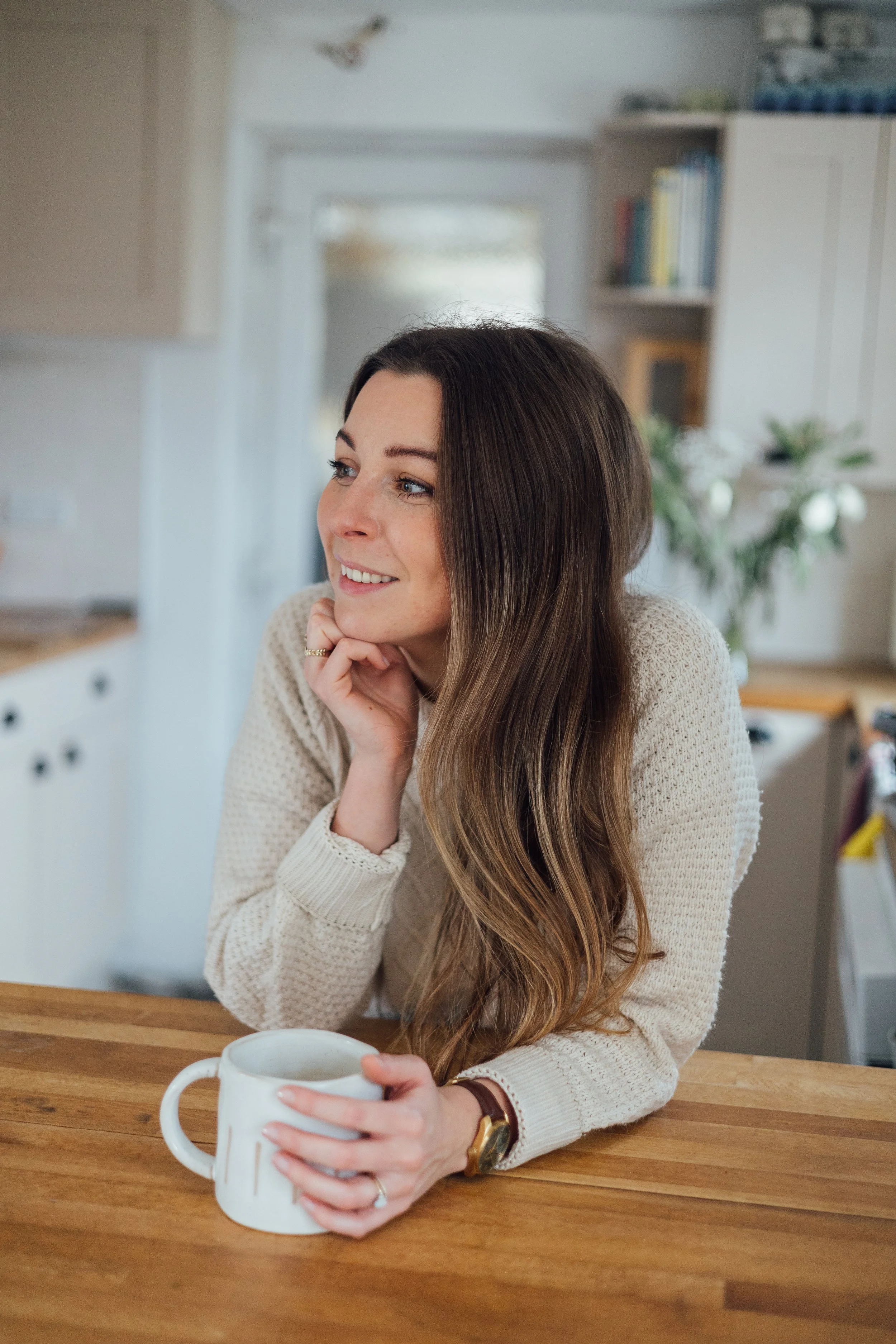 A woman with long brown hair, wearing a beige sweater and a watch, sits at a wooden table holding a white ceramic mug with both hands, smiling and looking to her left in a cozy kitchen.