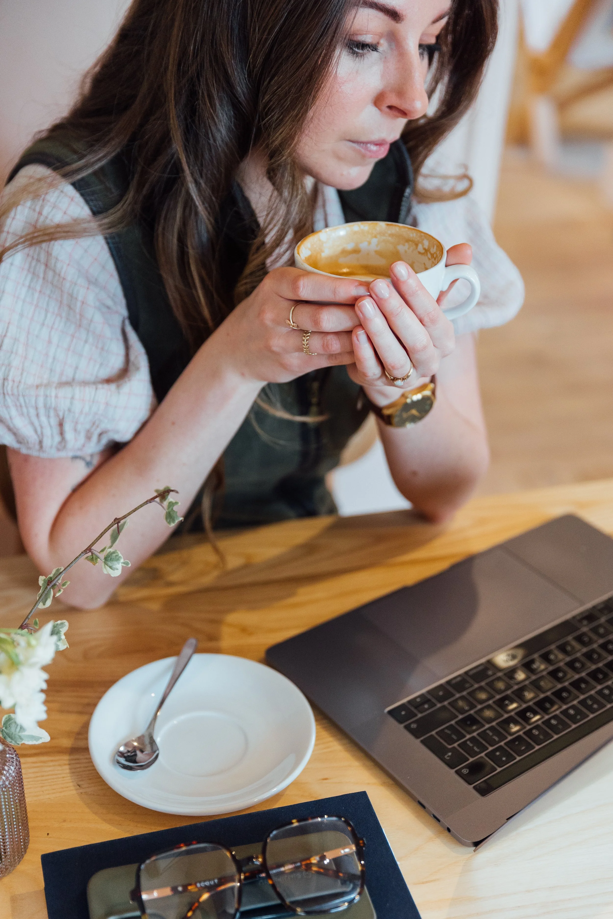 A woman with long brown hair sitting at a wooden table, holding a white mug, with various items on the table including a laptop, a white saucer, a spoon, a pair of glasses, and a small plant.