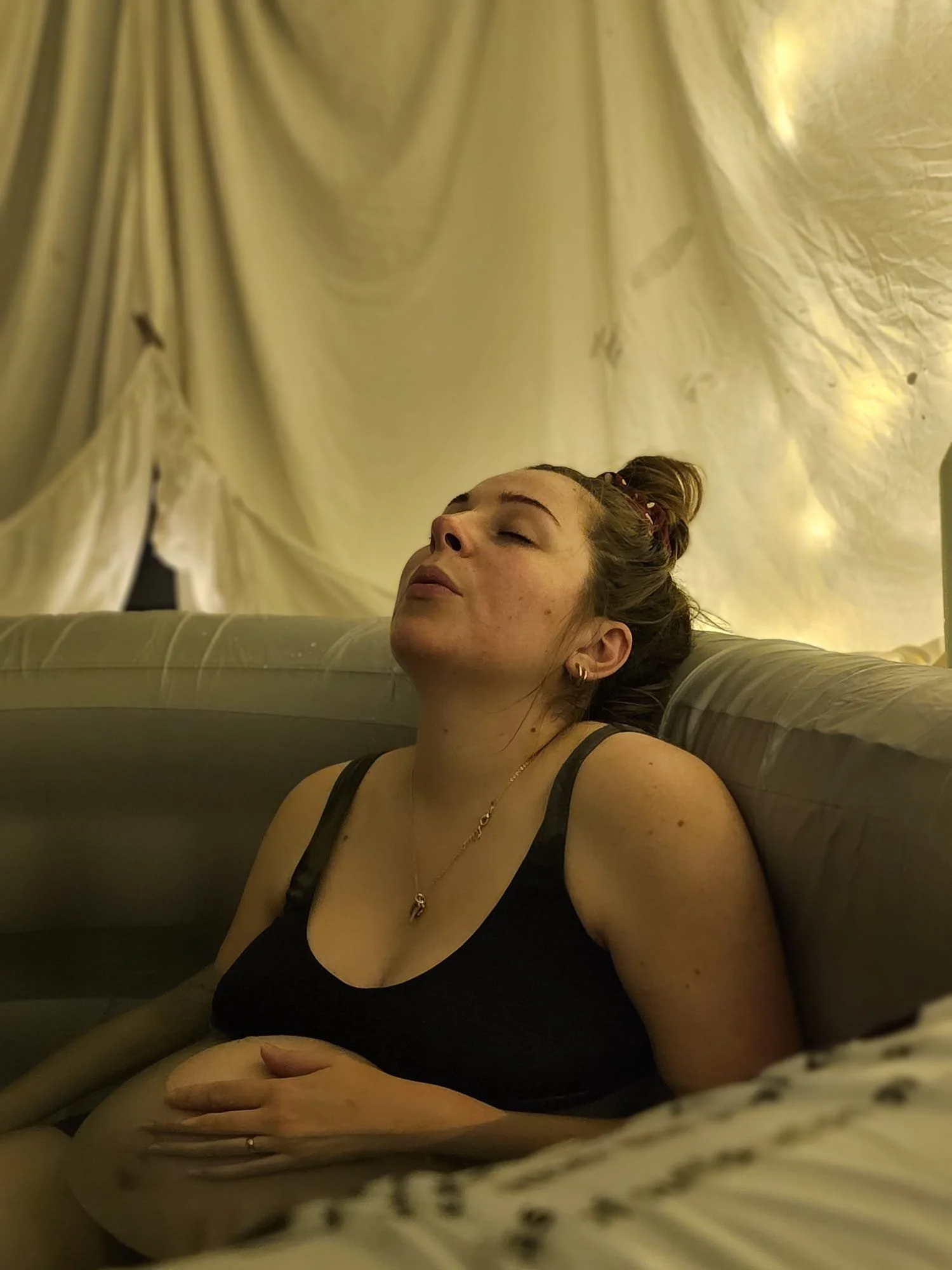 A woman with brown hair tied in a bun, closed eyes, and wearing a black tank top, appears to be resting or relaxing on a couch with her hand on her stomach, in front of a yellowish curtain.