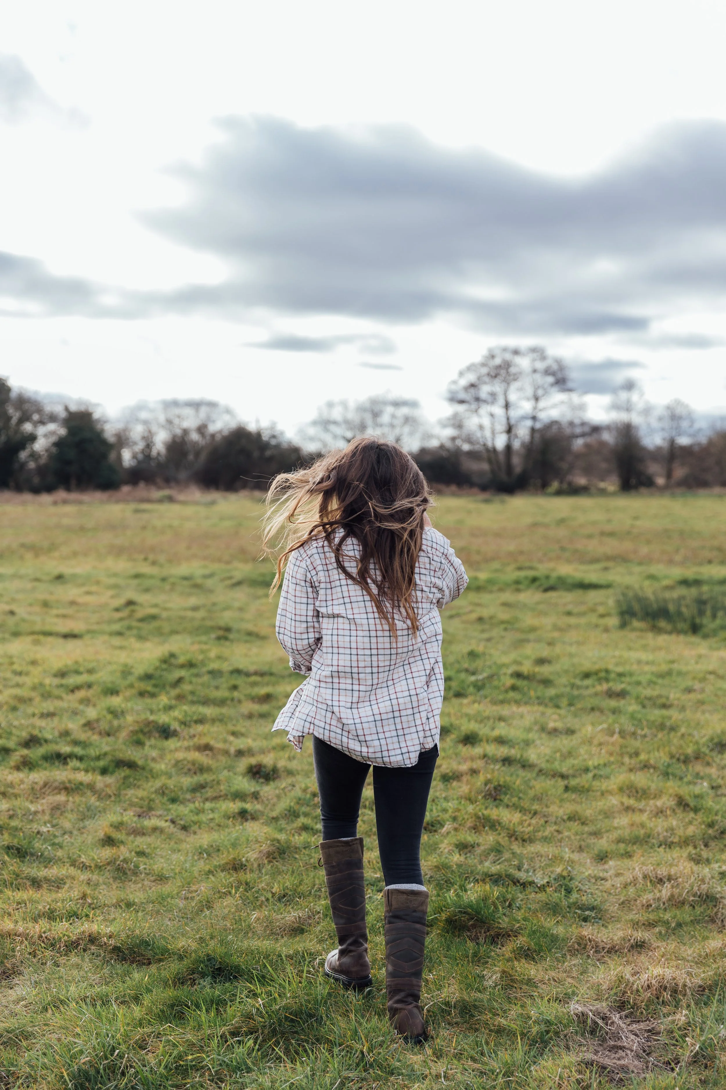 A girl with long, wavy hair wearing a checkered shirt, black pants, and tall brown boots walking in a grassy field during daytime with overcast sky and trees in the background.