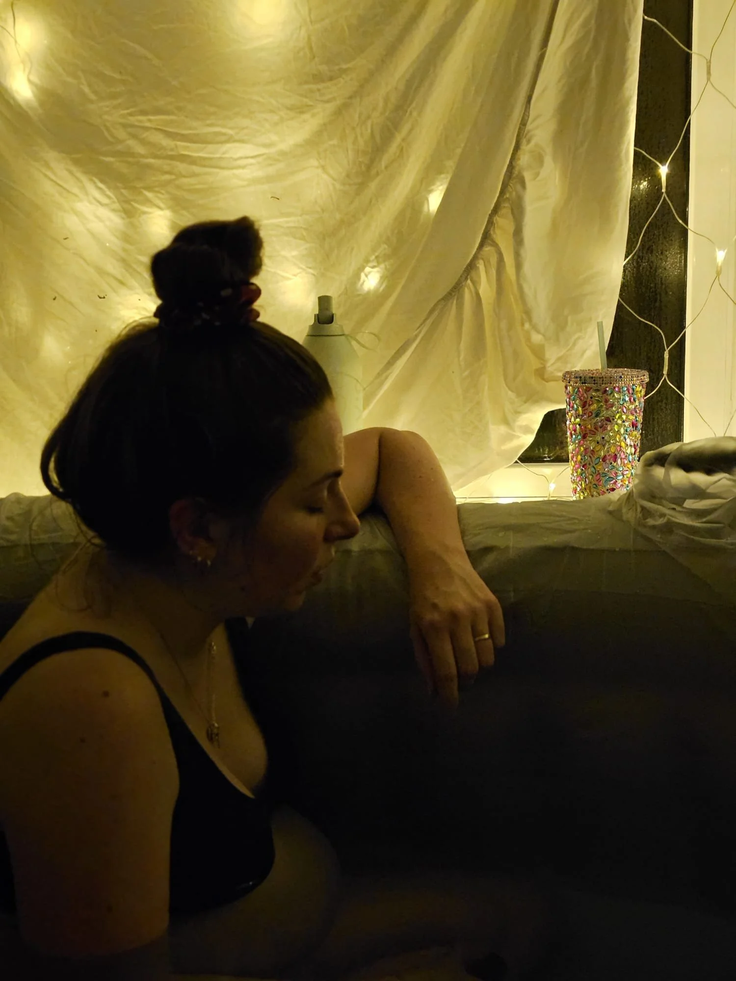 A woman with dark hair in a bun, wearing a black top, lying with closed eyes on a dark surface. Behind her, a beige curtain with fairy lights is draped, and a colorful, beaded cup with a straw is on a surface next to her.