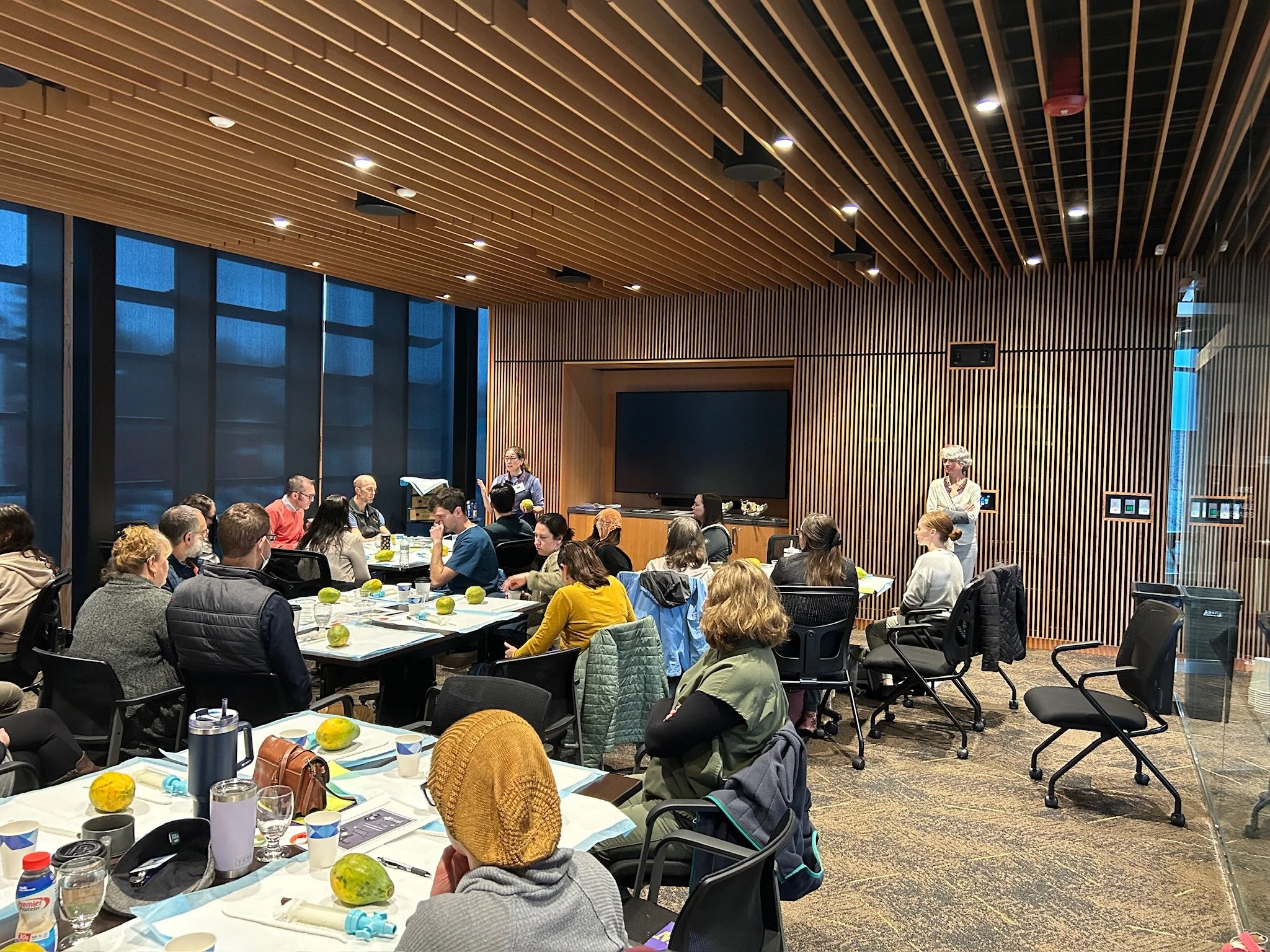 Multiple people seated at round tables in a conference room, with a presenter standing near a large television screen on a wood-paneled wall. The tables have cups, water bottles, and green fruits. The room has a ceiling with wooden slats and multiple ceiling lights, and large windows along one side.