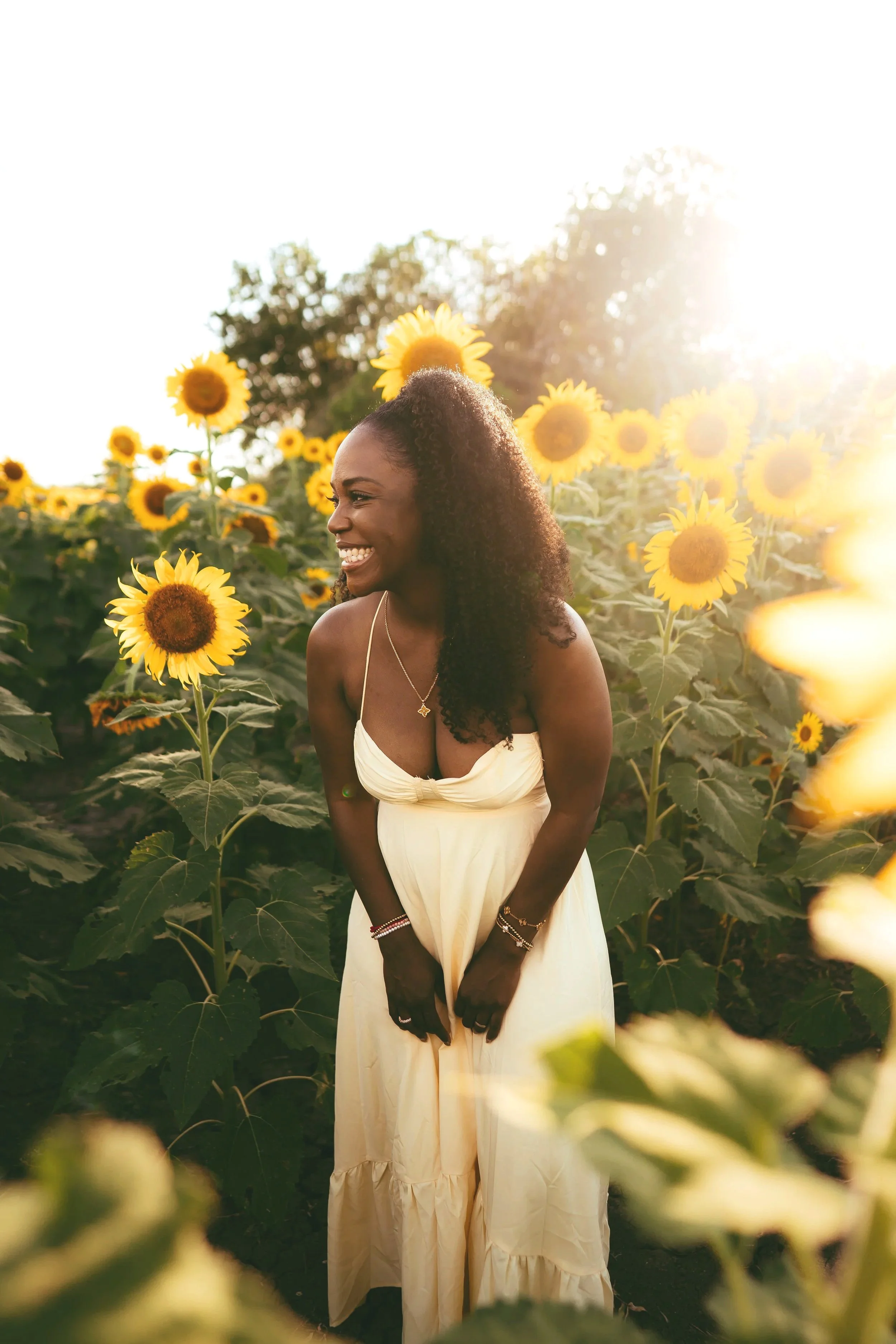 A woman standing in a sunflower field during sunset, smiling and looking to her left, wearing a white dress and jewelry.