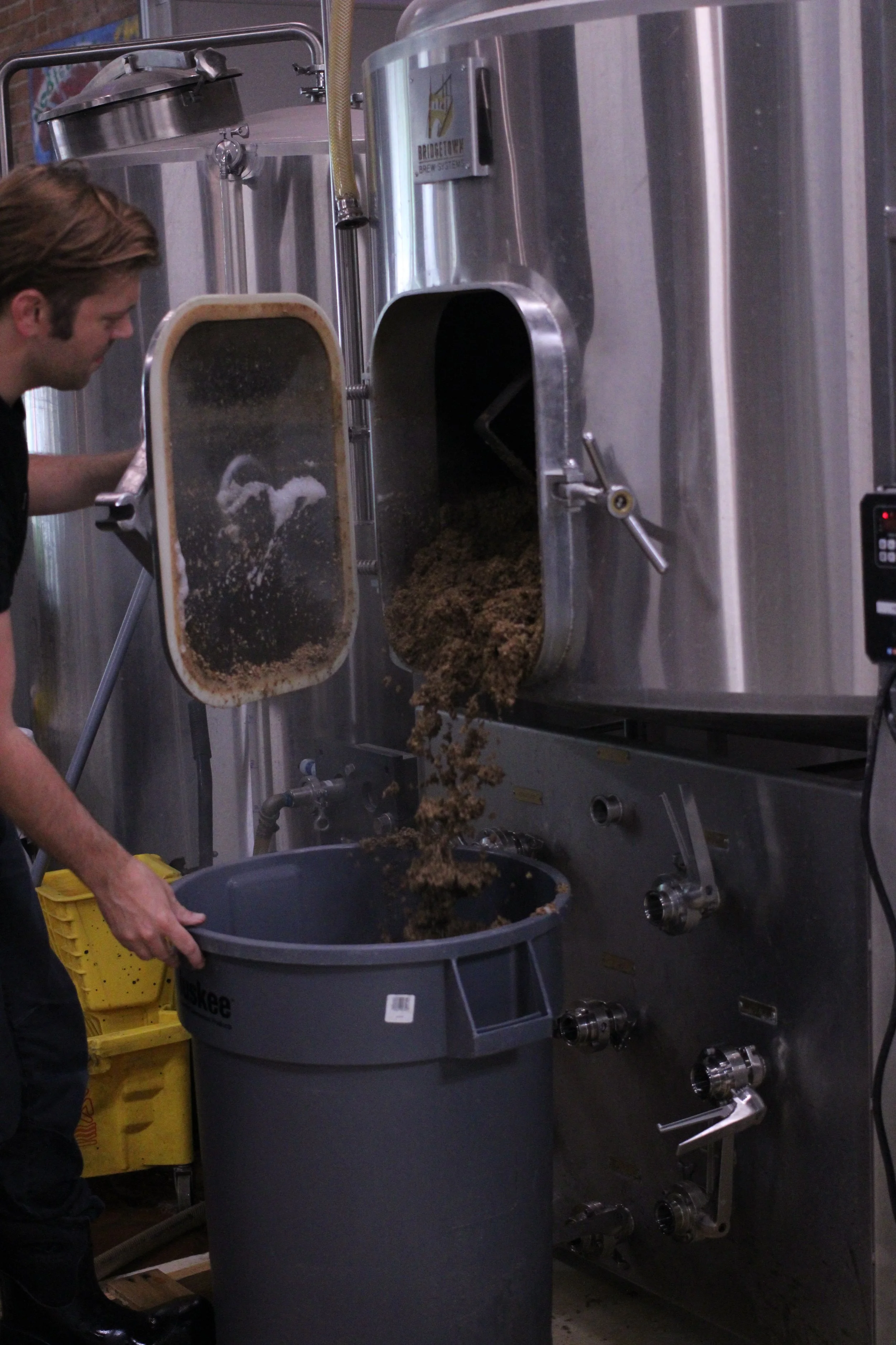 A person transfers ground meat from a large industrial meat grinder into a gray plastic tub. The meat grinder, made of stainless steel, has a large opening and a spout from which the meat is falling into the tub. The scene appears to be in a commercial kitchen or meat processing facility.