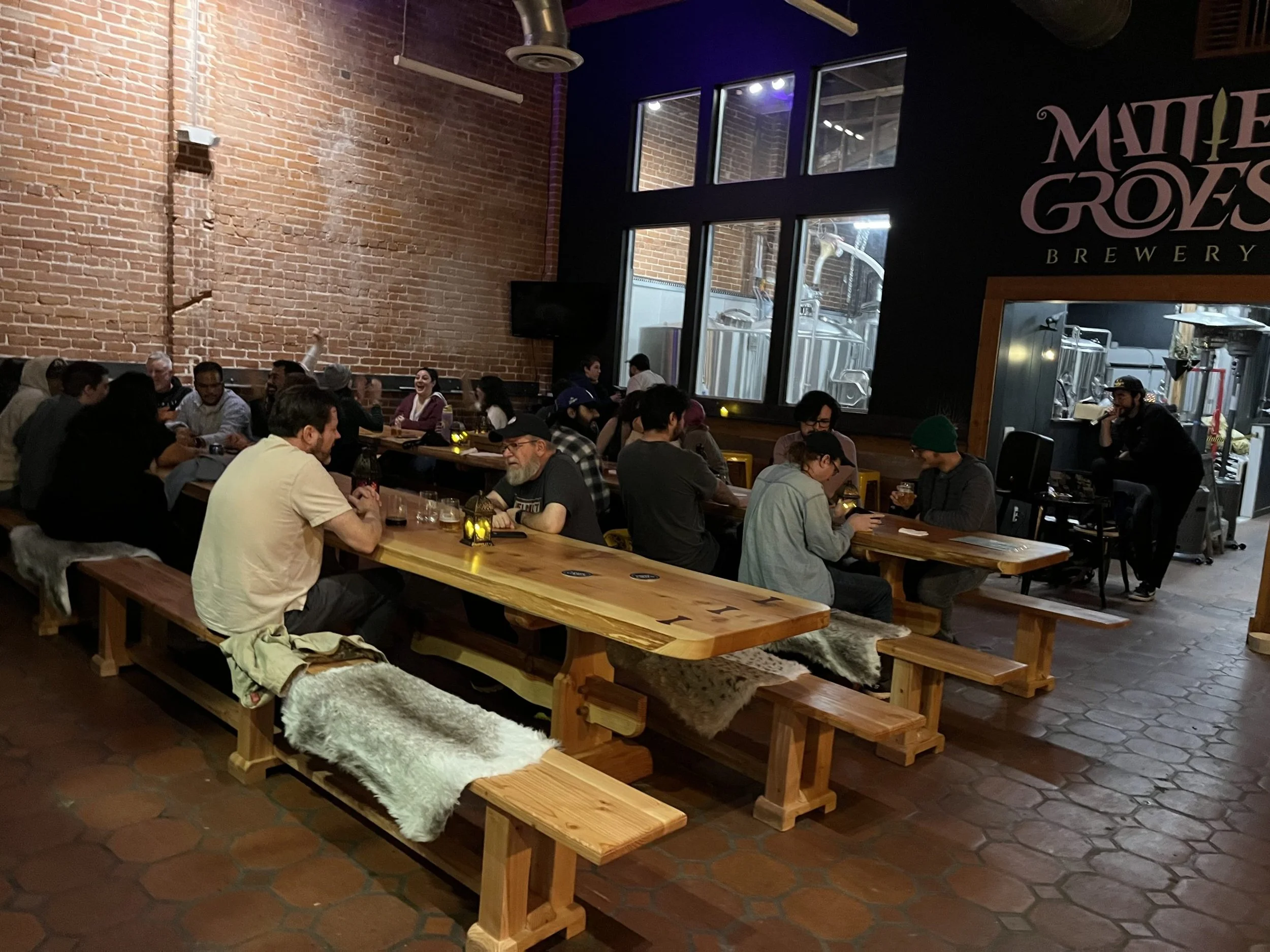 Inside of Matinee Grows Brewery with a large group of people seated at long wooden tables, exposed brick walls, and brewery tanks visible through large windows.