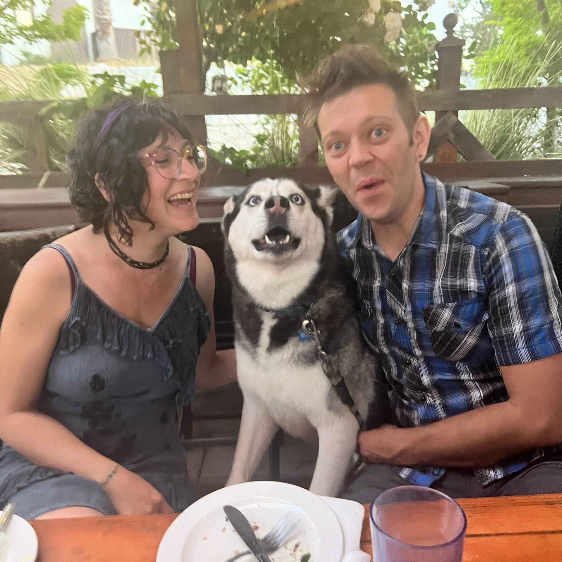 Two women and a husky dog sitting at a table outdoors, smiling and enjoying a meal. The woman on the left has curly dark hair, glasses, and is wearing a dark dress, while the man on the right has short brown hair, a plaid shirt, and earrings. The background features greenery and a wooden fence.