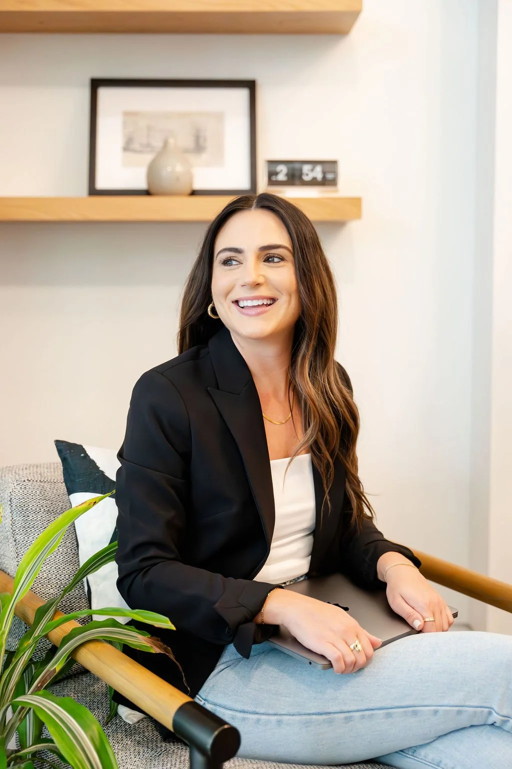A woman with long brown hair, wearing a black blazer and white top, sits on a gray couch with a smile, holding a laptop, with a plant nearby and a wooden shelf with decor behind her.
