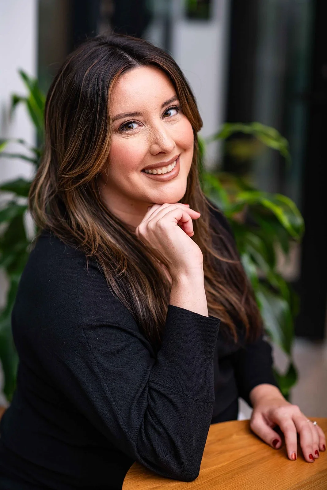 A woman with long brown hair smiling while resting her chin on her hand, seated at a wooden table with a background of green plants.