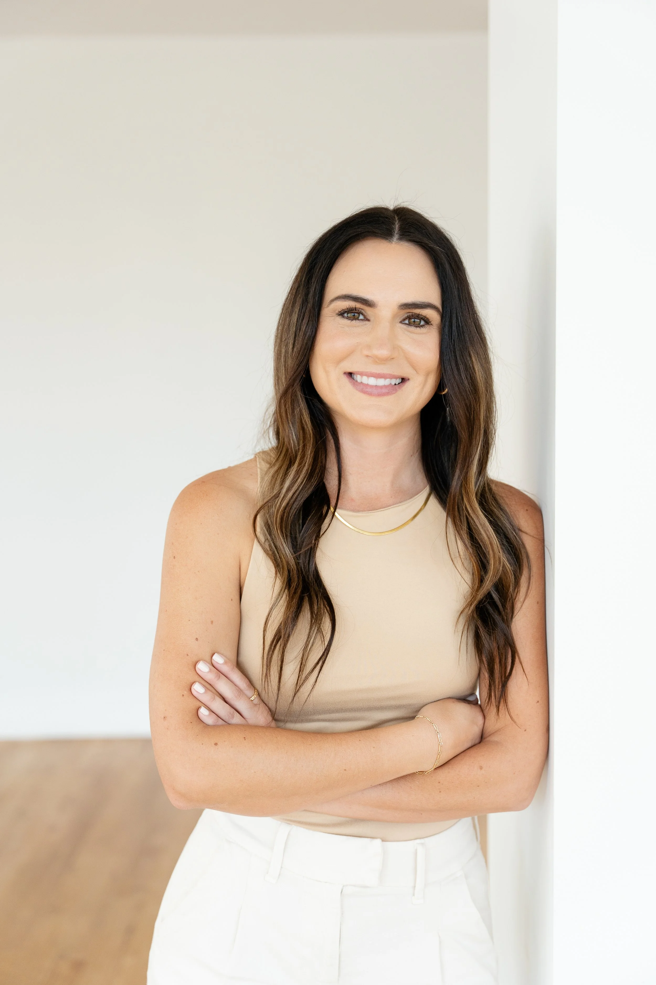 A woman with long wavy brown hair, wearing a beige sleeveless top and white pants, smiling and standing with her arms crossed in front of a white wall.