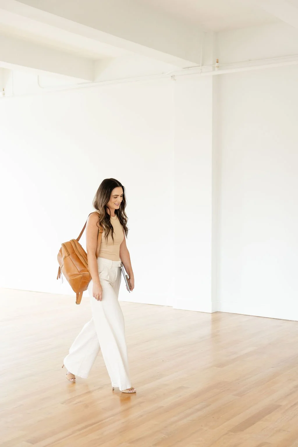 Woman with long dark hair walking in a bright white room, carrying a brown leather backpack on her back and a book in her hand, wearing white wide-leg pants and a beige sleeveless top.