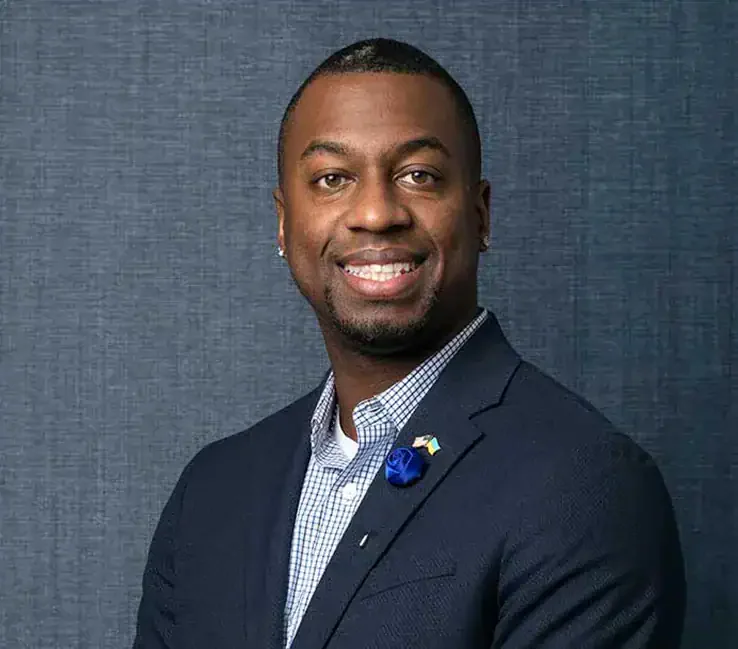 A smiling African American man in a navy suit with a checkered shirt, standing against a dark textured background.