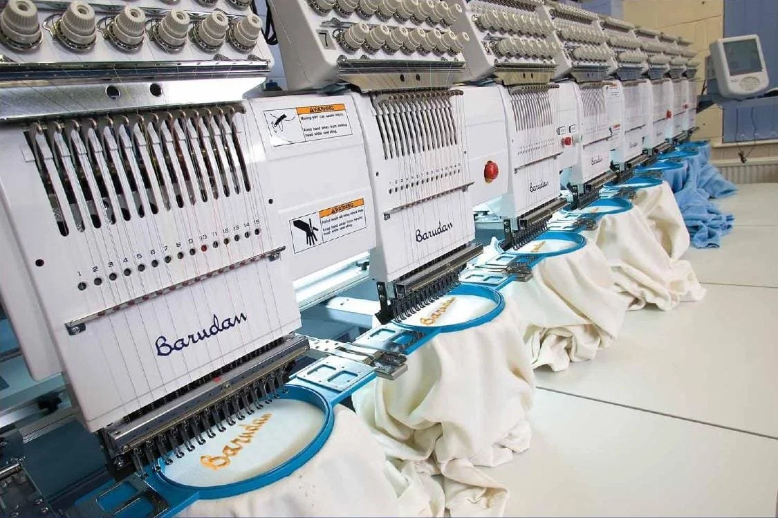 Multiple embroidered embroidery machines stitching the word 'Barudan' onto fabric in a factory setting.