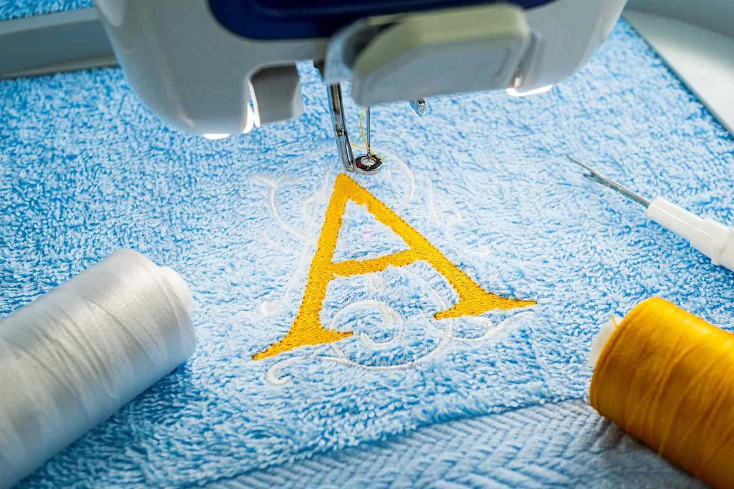 Close-up of embroidery machine stitching the word 'AR' in yellow on a blue terry cloth towel, with spools of white and yellow thread nearby.