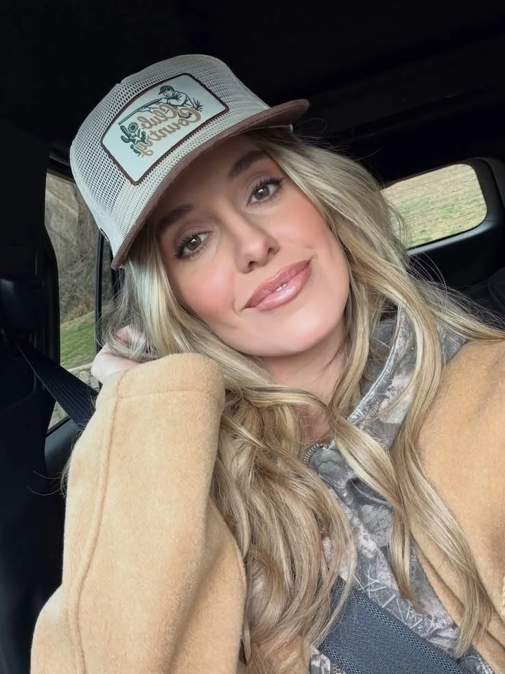 Young woman with long blonde hair wearing a camouflage hoodie, tan coat, and a mesh trucker cap with a logo and text, sitting inside a vehicle.