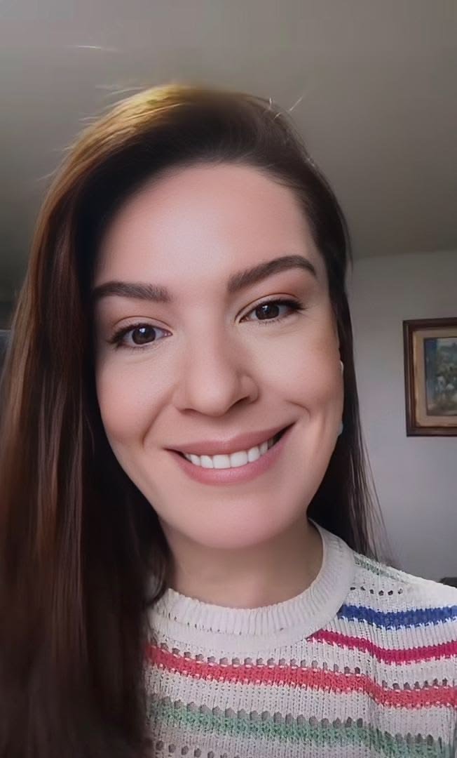 Close-up of a woman with long brown hair, smiling in a room with a painting on the wall behind her, wearing a striped sweater.