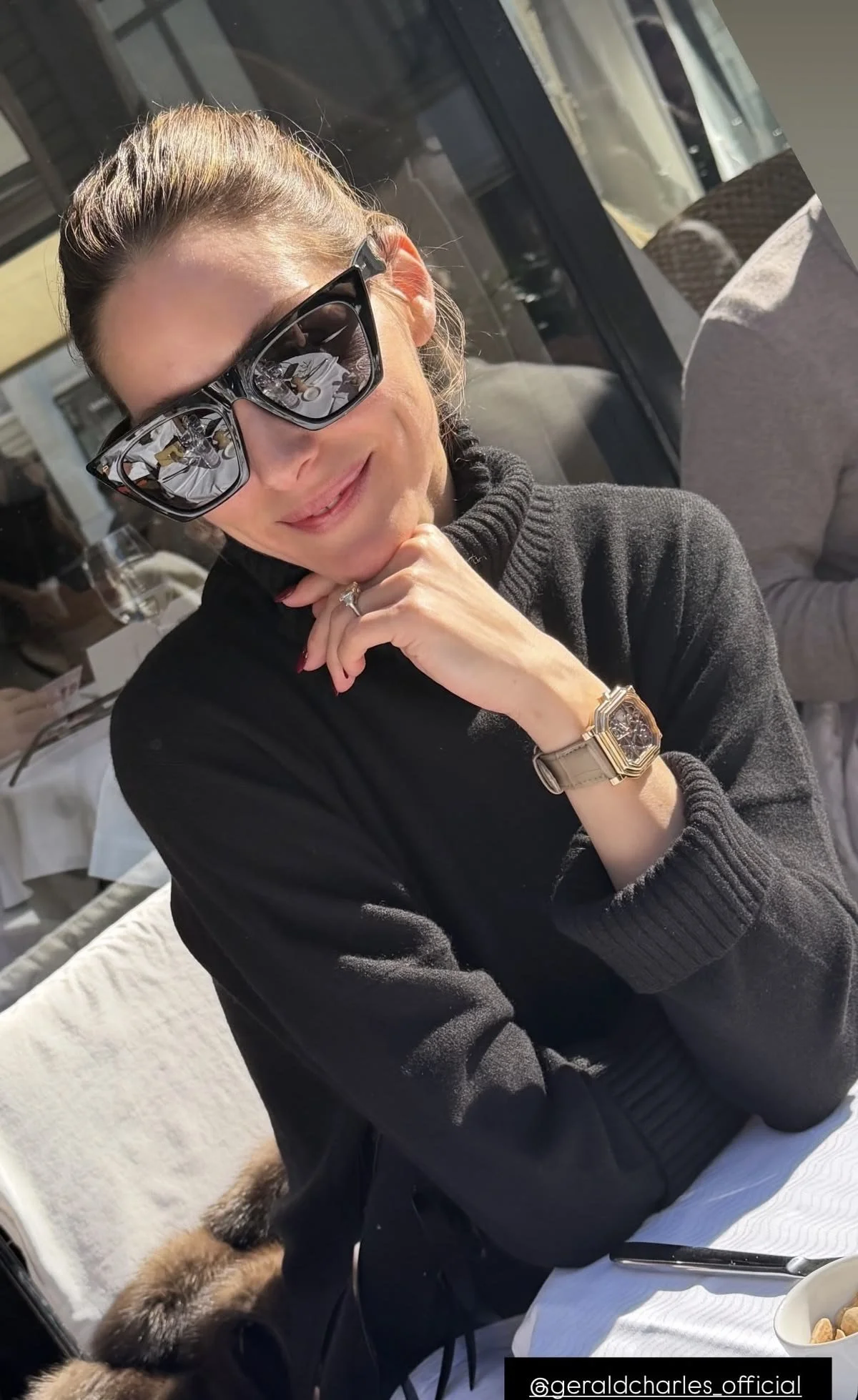 A woman sitting at an outdoor restaurant table with sunglasses, a watch, and a ring, resting her chin on her hand. She is dressed in a black turtleneck sweater, and part of a person in a gray shirt and a dog with fur are visible nearby.