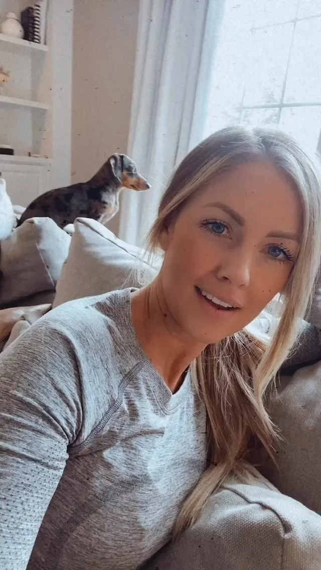 A woman with long blonde hair and blue eyes taking a selfie inside a living room with a dog in the background.