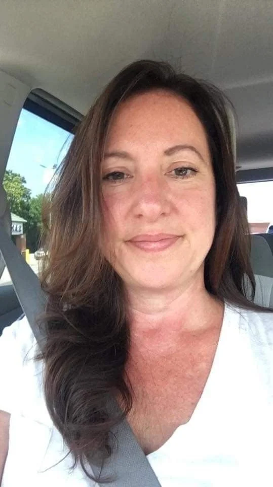 A woman with long brown hair taking a selfie inside a vehicle, with trees and buildings visible outside the window.