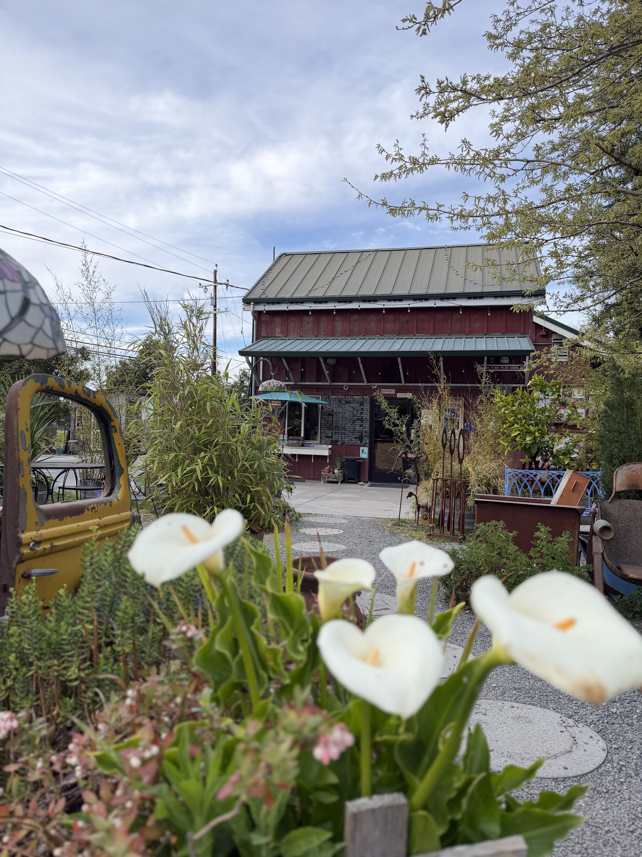 A red barn with a metal roof in a garden setting, surrounded by various plants and flowers, with a clear pathway leading to the entrance and some outdoor furniture visible.