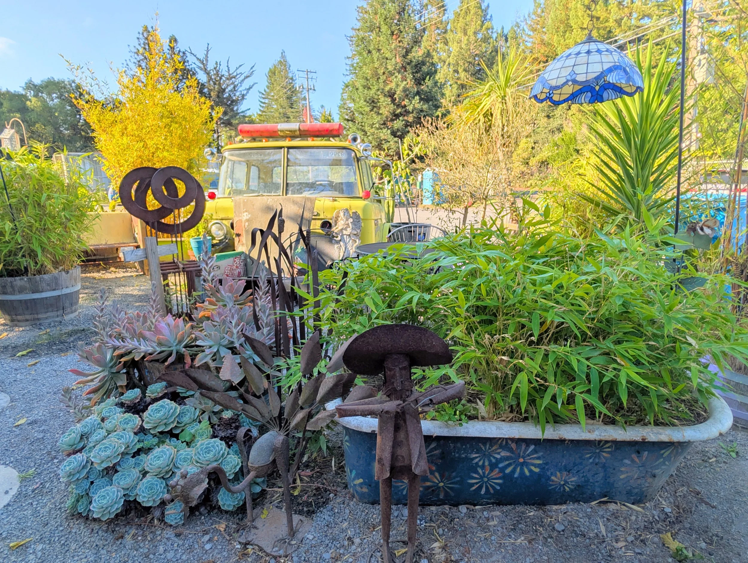 A garden scene with various potted plants including succulents, a large painted ceramic pot, a rusty metal sculpture, and an old yellow fire truck in the background. There is also a stained glass lamp hanging among the greenery.