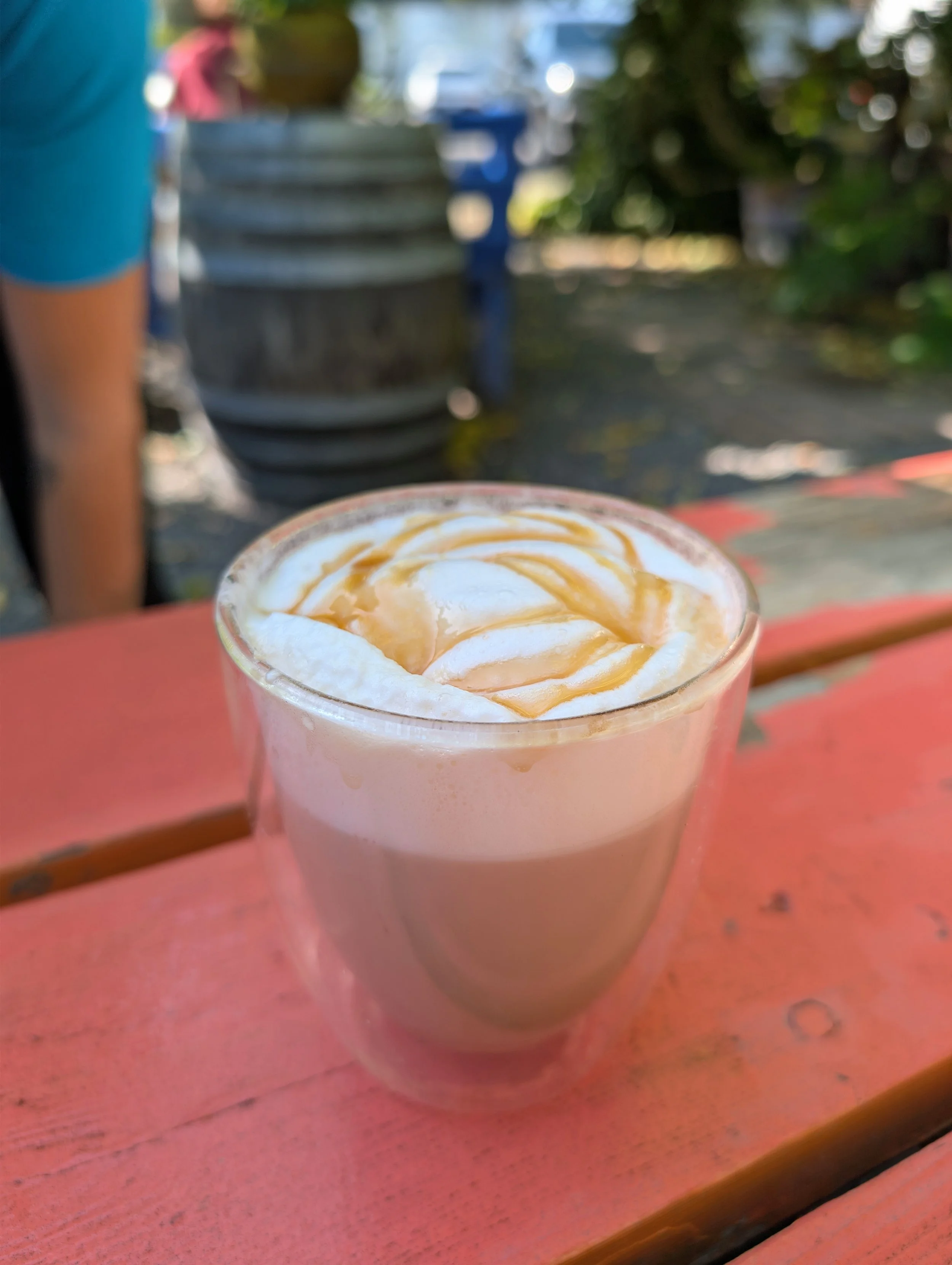 A caramel-flavored coffee drink topped with whipped cream and caramel drizzle in a clear plastic cup, on a pinkish-orange wooden table outdoors.
