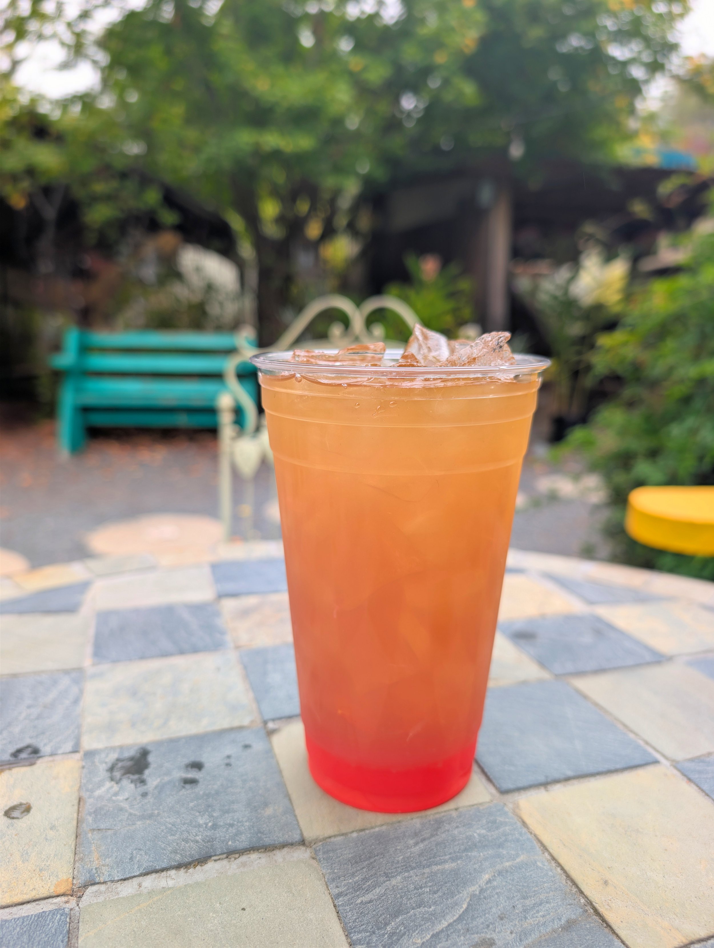 A cup filled with a pinkish-orange mate and ice cubes, placed on a checkered table outdoors with a blue bench, trees, and a wooden structure in the blurred background.
