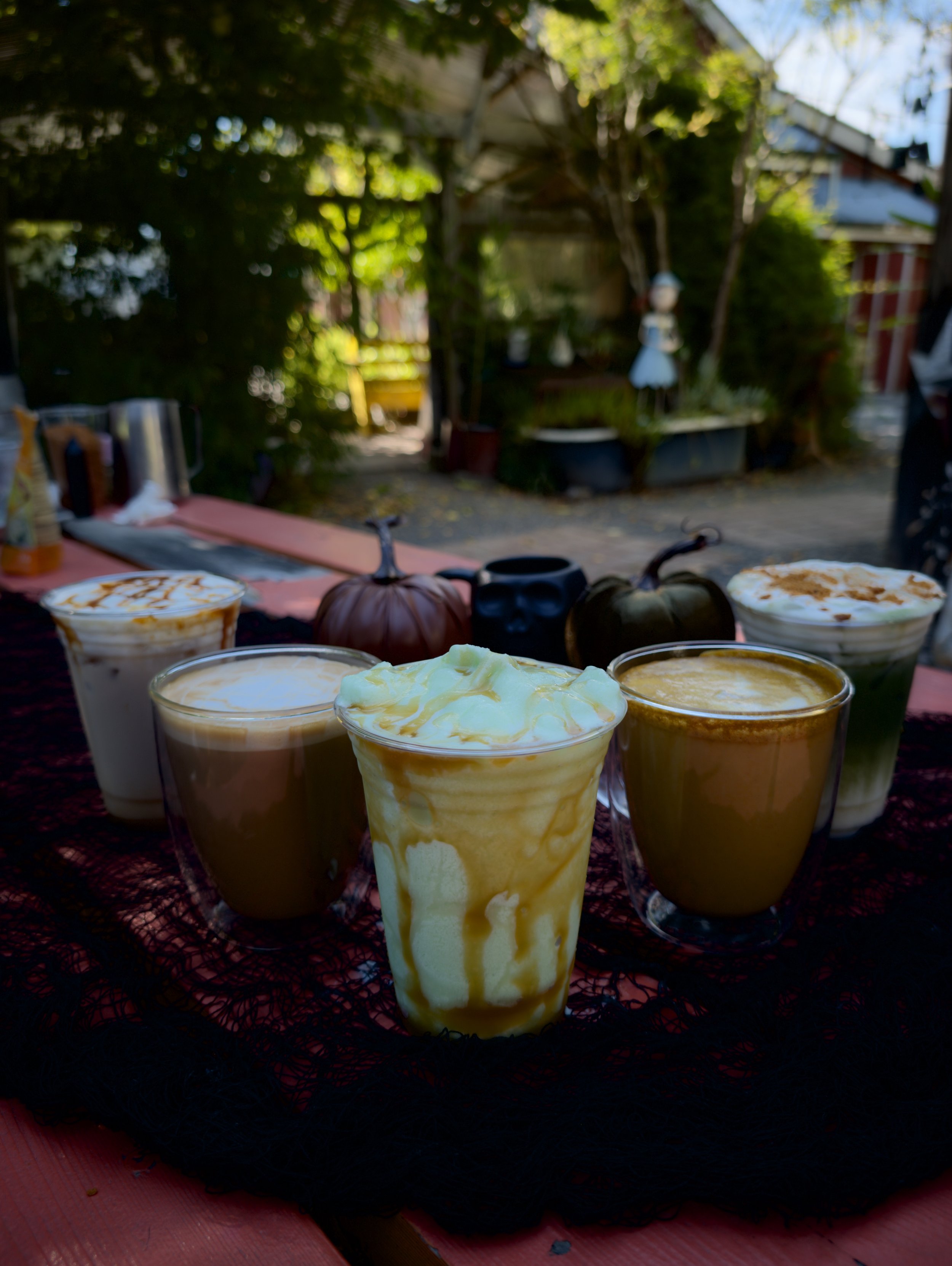 Several tall glasses filled with caramel-colored drinks topped with whipped cream, placed on a red-patterned cloth. The background shows an outdoor setting with greenery, trees, and a fence.