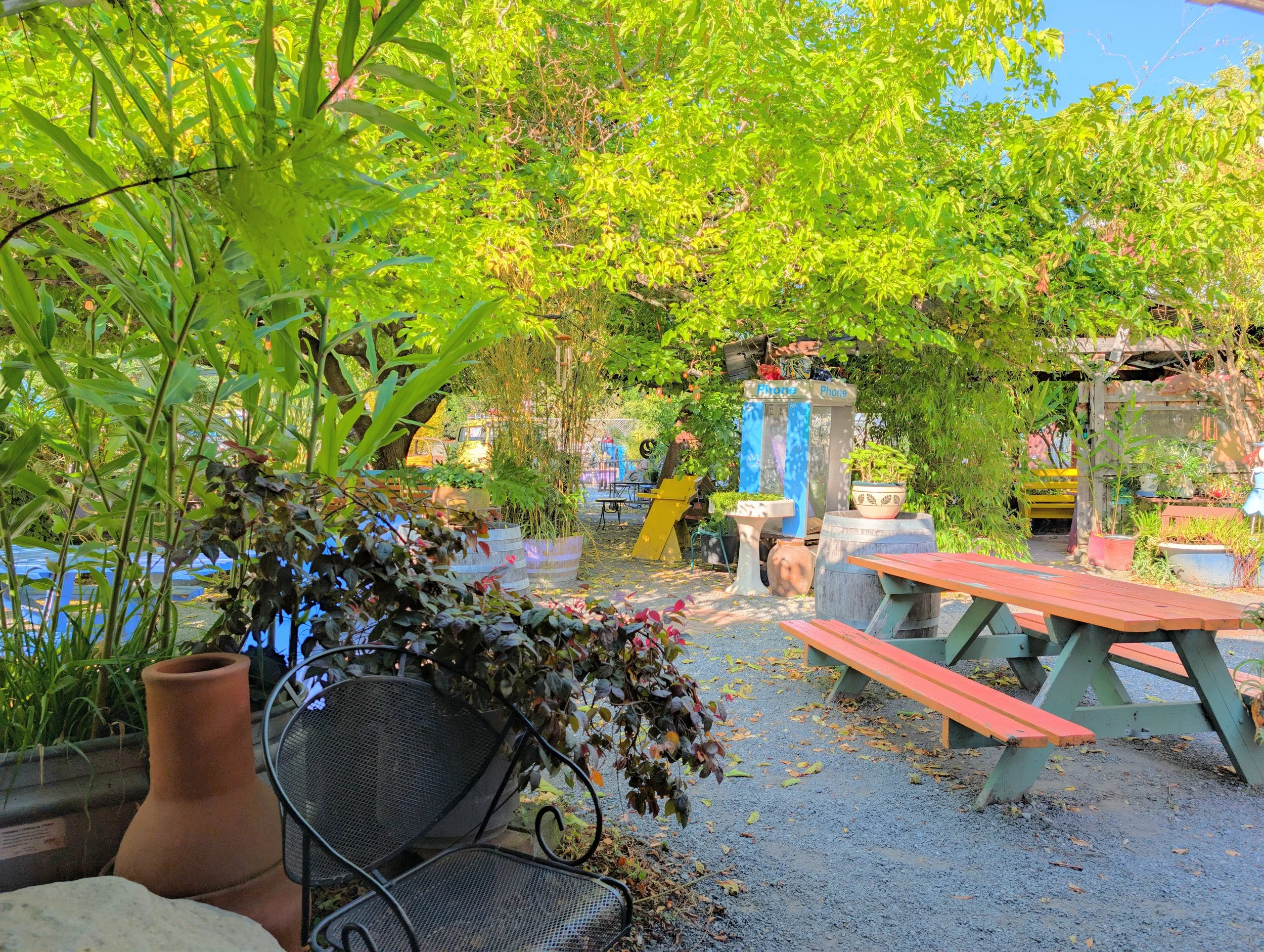 An outdoor cafe area with wooden picnic tables painted in pastel pink and green, surrounded by lush green plants and large trees providing shade, with a blue phone booth and some chairs in the background.