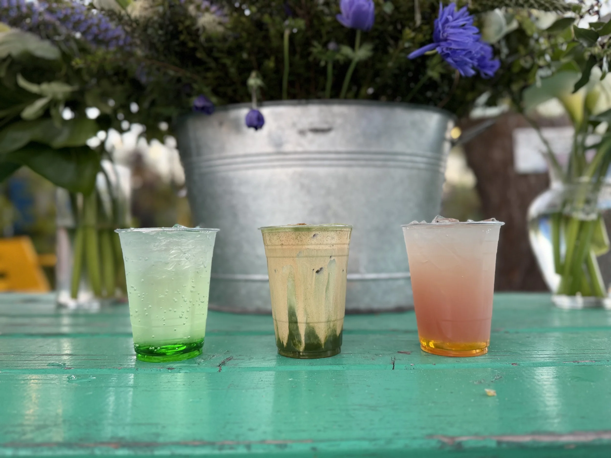 Colorful beverages in clear cups placed on a green wooden table, with a planter full of purple and white flowers in the background.