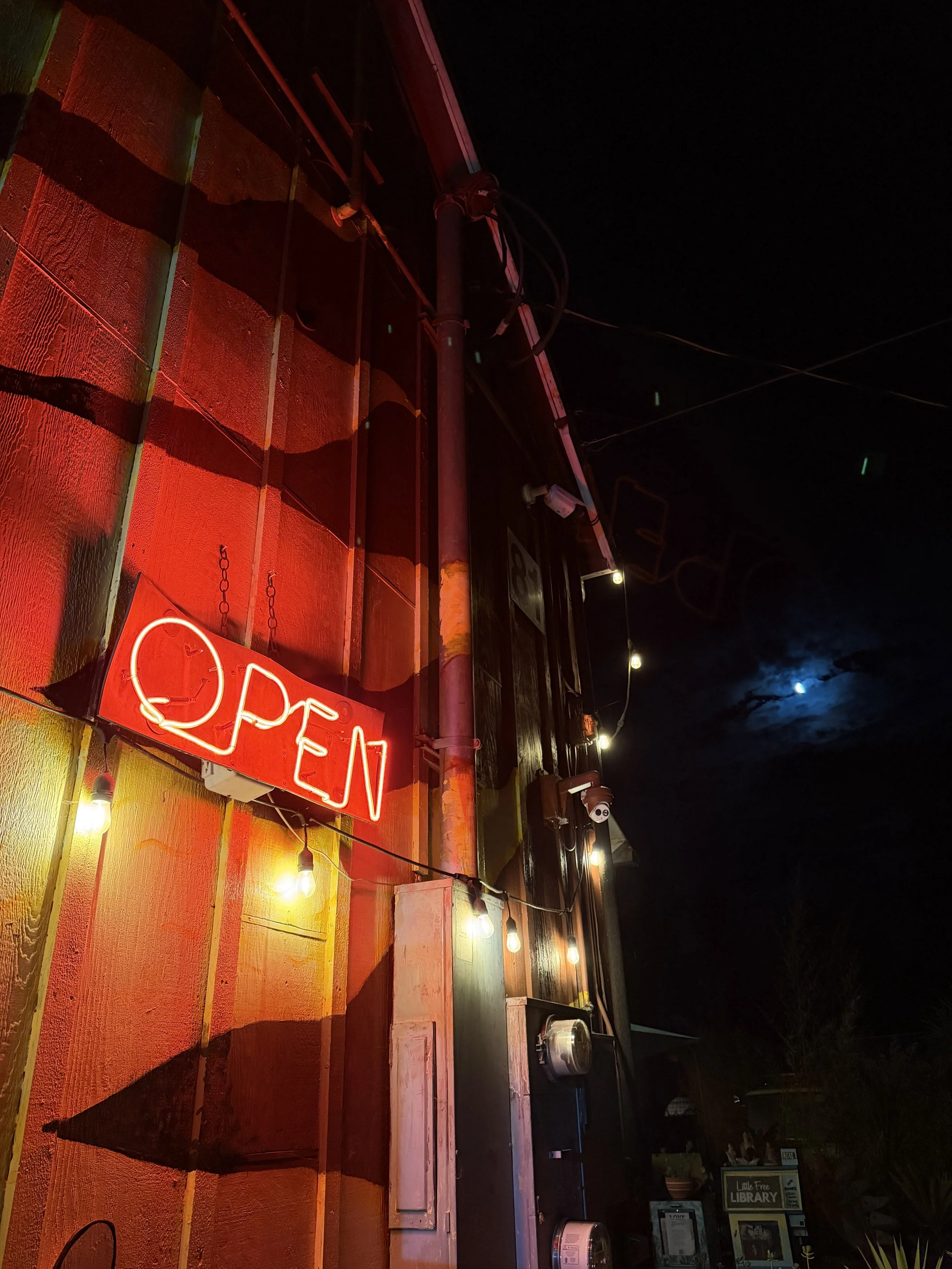 Night scene showing a building with a red neon 'OPEN' sign, string lights hanging along the wall, and a partly visible sign for a little free library against a dark sky with the moon.